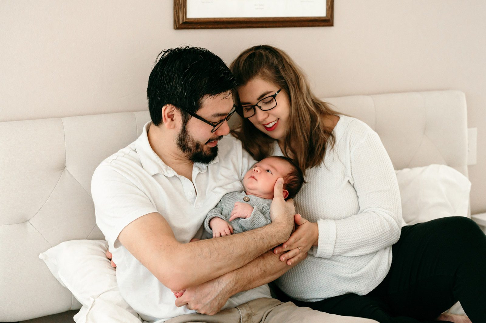 new parents sitting on their bed and smiling down at their baby boy during an in home newborn photoshoot