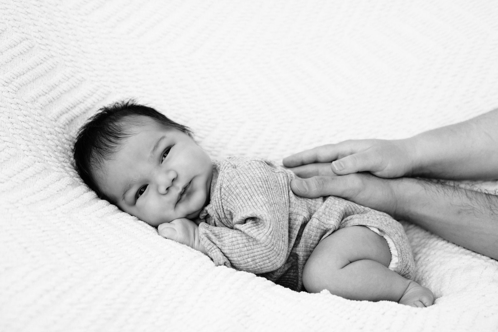 a black and white picture of a newborn baby boy laying on a white backdrop with his parents resting their hands on his back during a newborn photoshoot