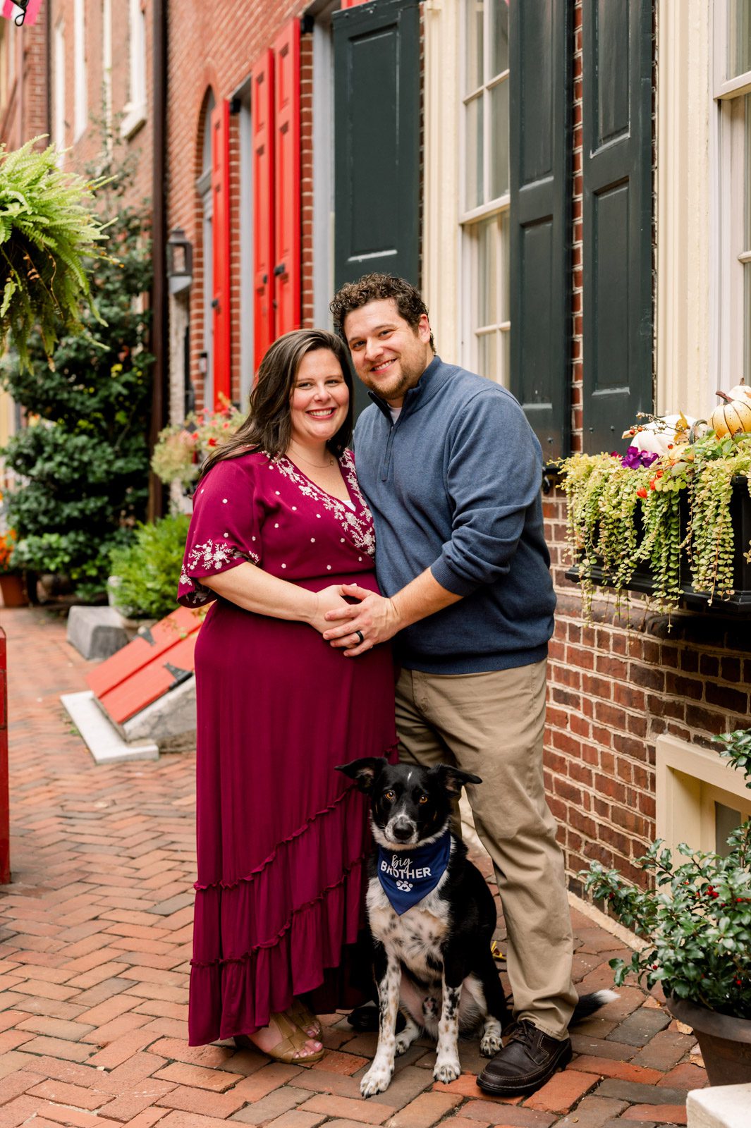 expecting parents and their pet dog standing in Elfreth's Alley and smiling at the camera during a Philadelphia maternity photoshoot