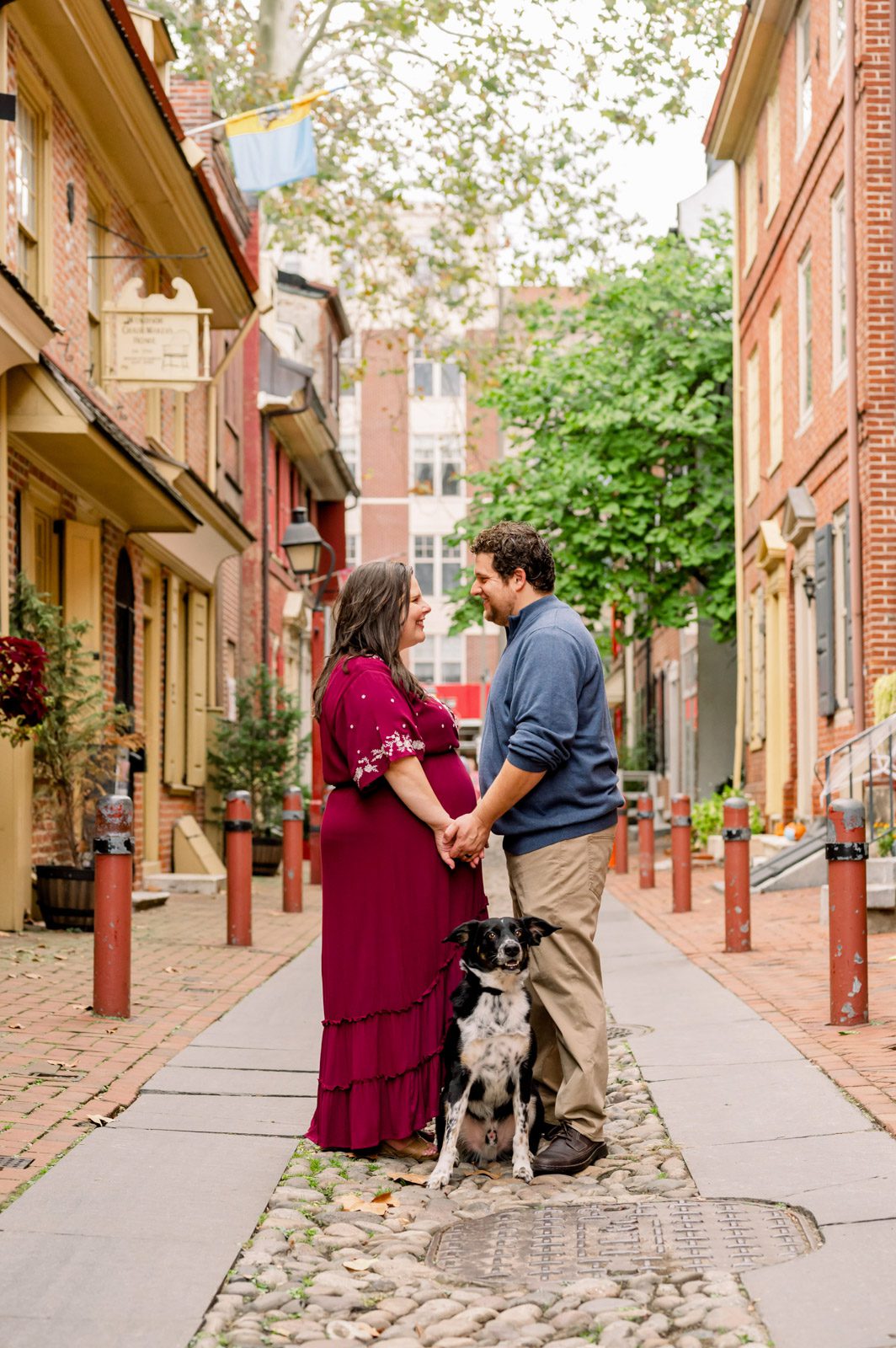 an expecting couple standing in Elfreth's Alley and smiling at each other with their pet dog at their feet during a Philadelphia maternity photoshoot