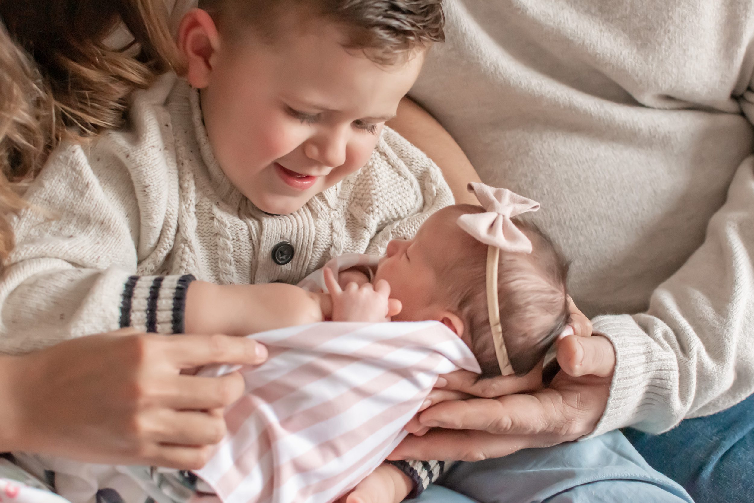 Boy sitting in his parents lap holding his newborn baby sister who is grabbing onto his finger during an in home newborn photography session