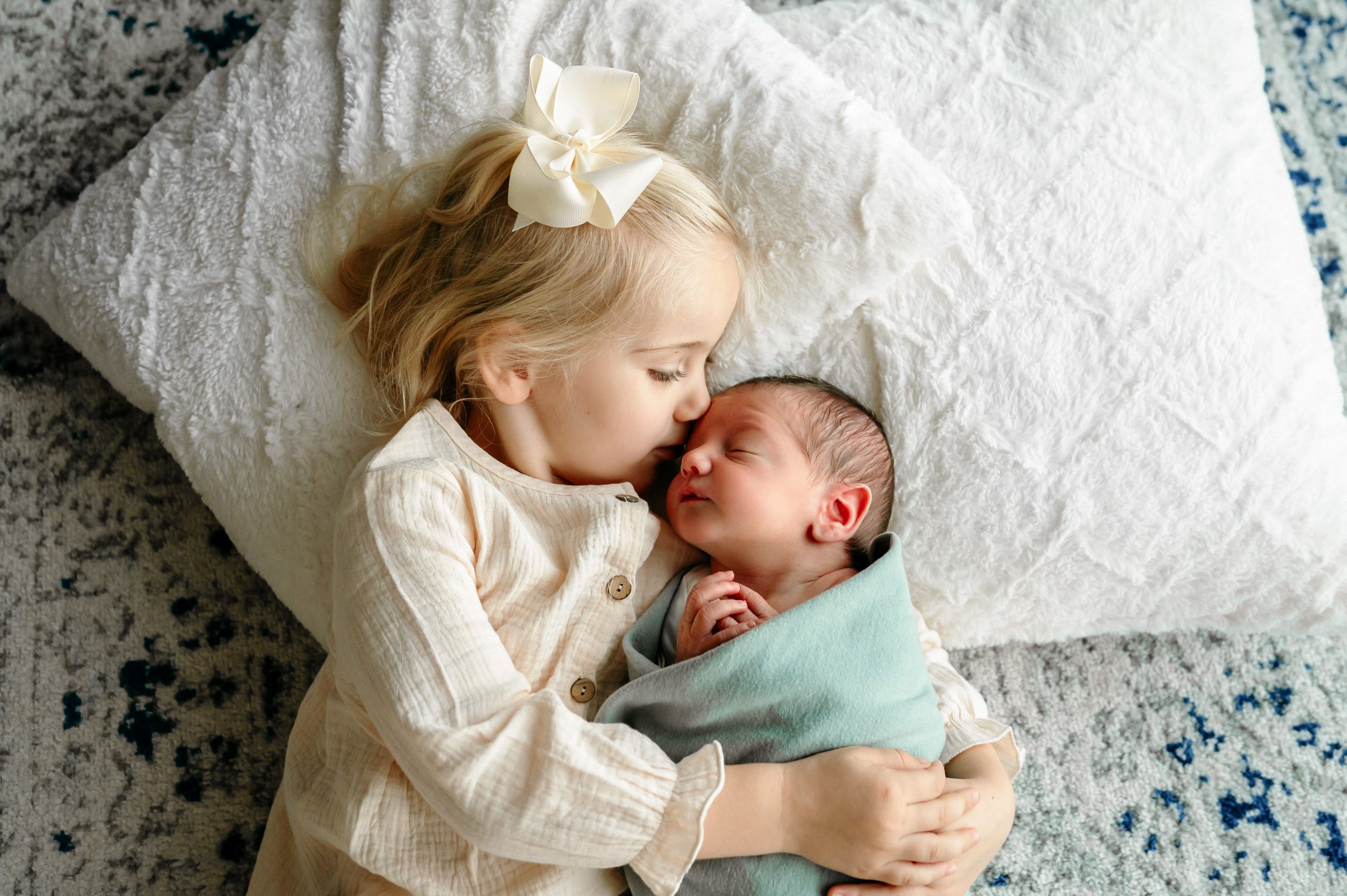 Older sister laying on pillows kissing her newborn baby brother on the forehead during an in home newborn photoshoot