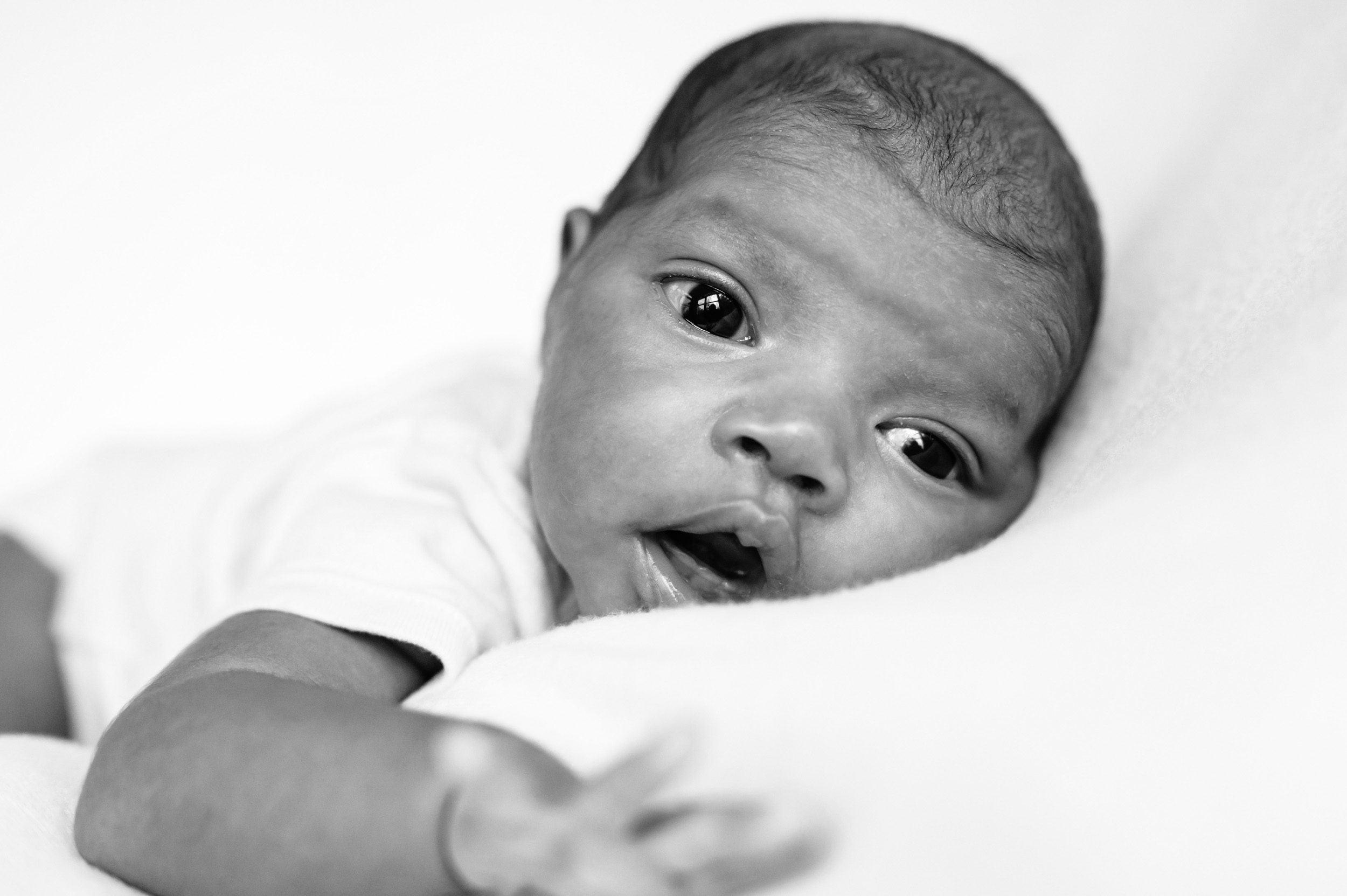 black and white image of a newborn baby girl on a white backdrop reaching out toward the camera during a newborn photography session