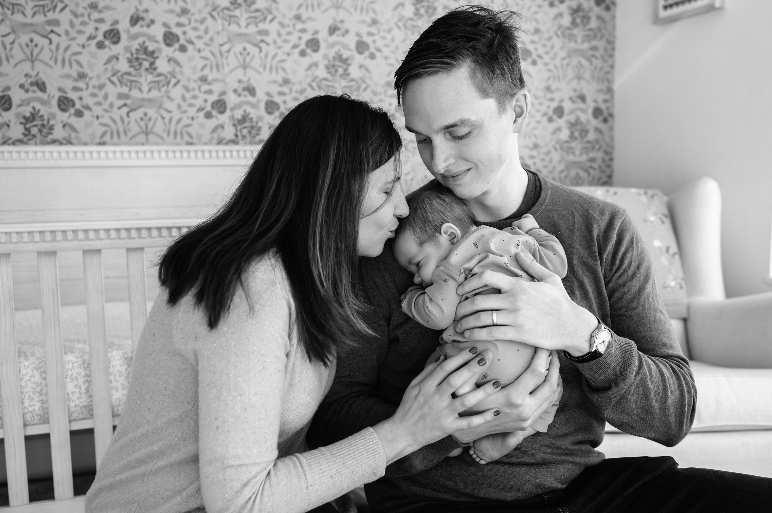 Black and white picture of a dad holding his newborn son while mom kisses him on the head during an in home newborn photography session