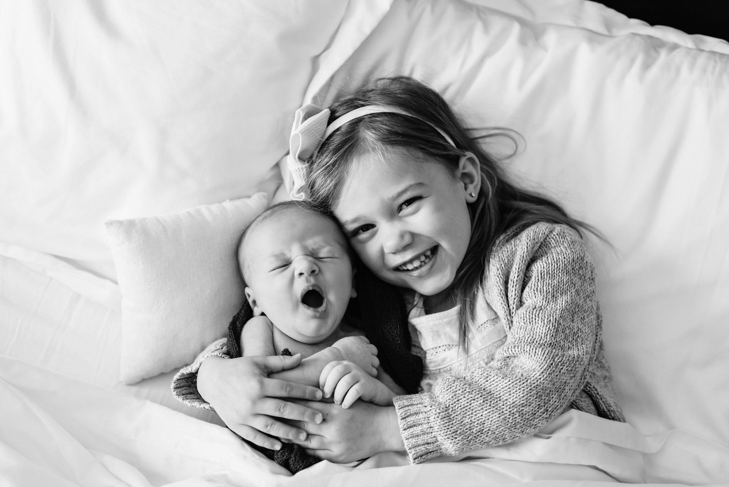 Black and white image of older sibling hugging her newborn baby brother and smiling up at the camera during an in home newborn photo session