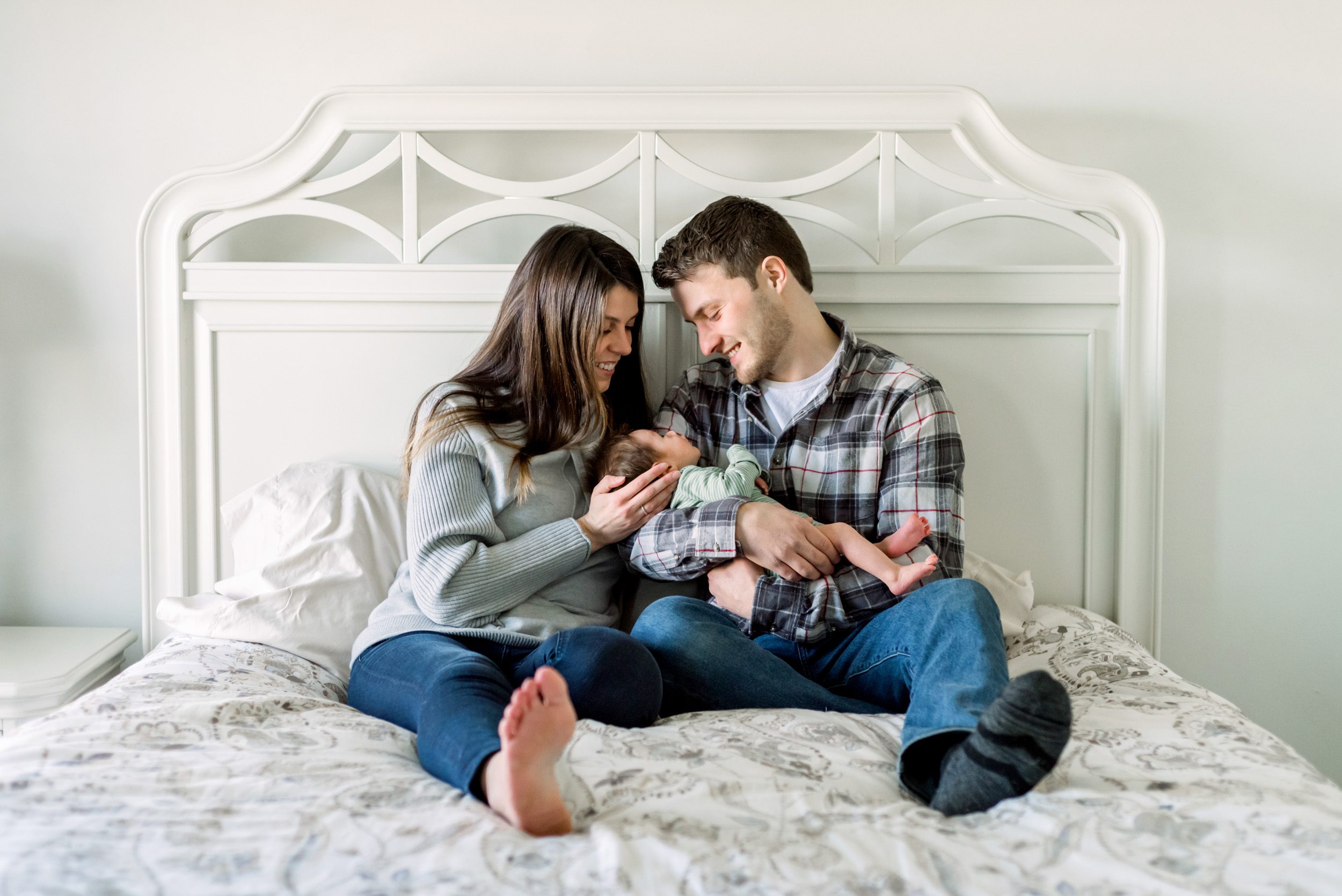 Newborn picture of parents sitting on a bed and smiling down at their baby son in their arms during an in home newborn photo session