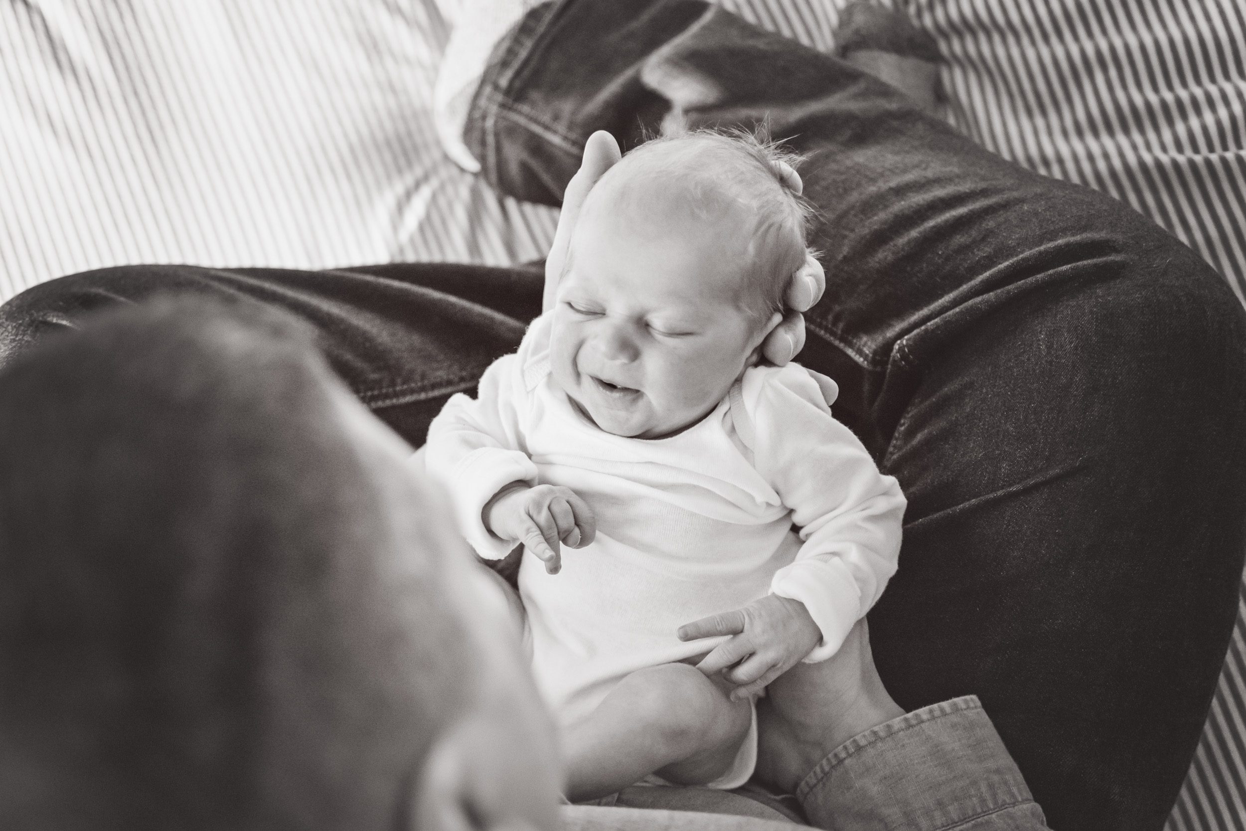 Black and white picture of a newborn boy sitting in his dad's lap smiling during a natural newborn photoshoot