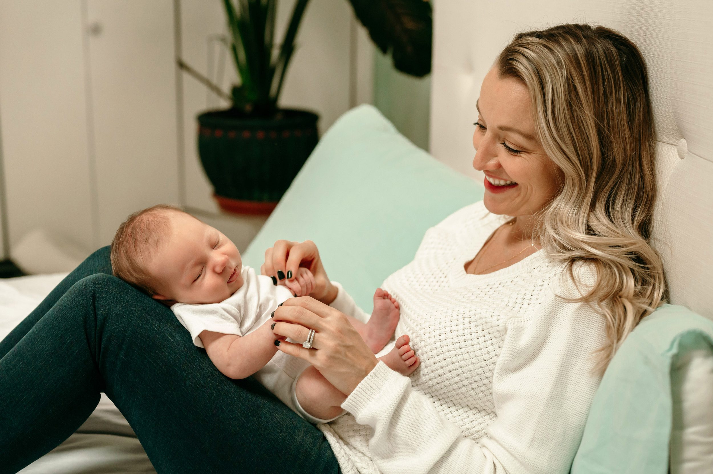 Newborn picture of a mom sitting on a bed with her baby boy laying on her legs and smiling at him during a lifestyle newborn photo session