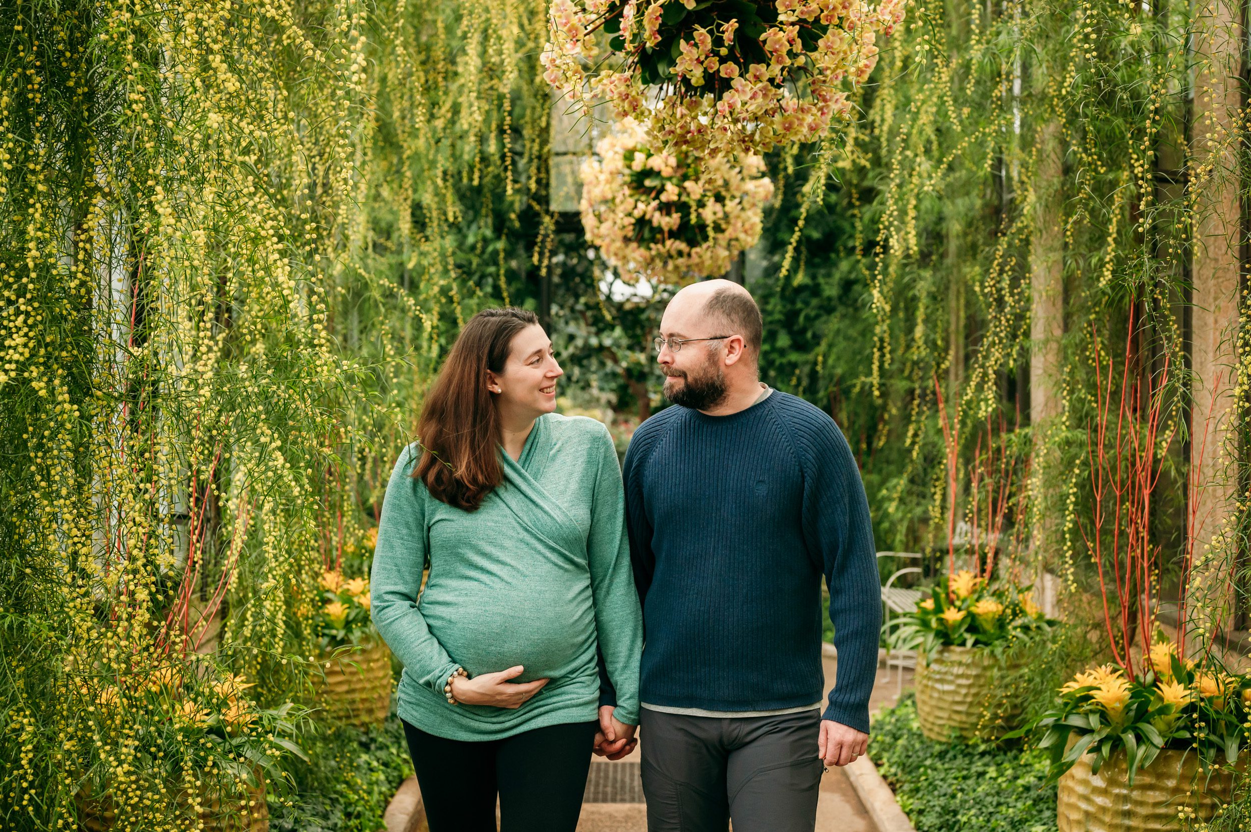 expecting parents holding hands and smiling at each other as they walk through a colorful greenhouse at a maternity photoshoot
