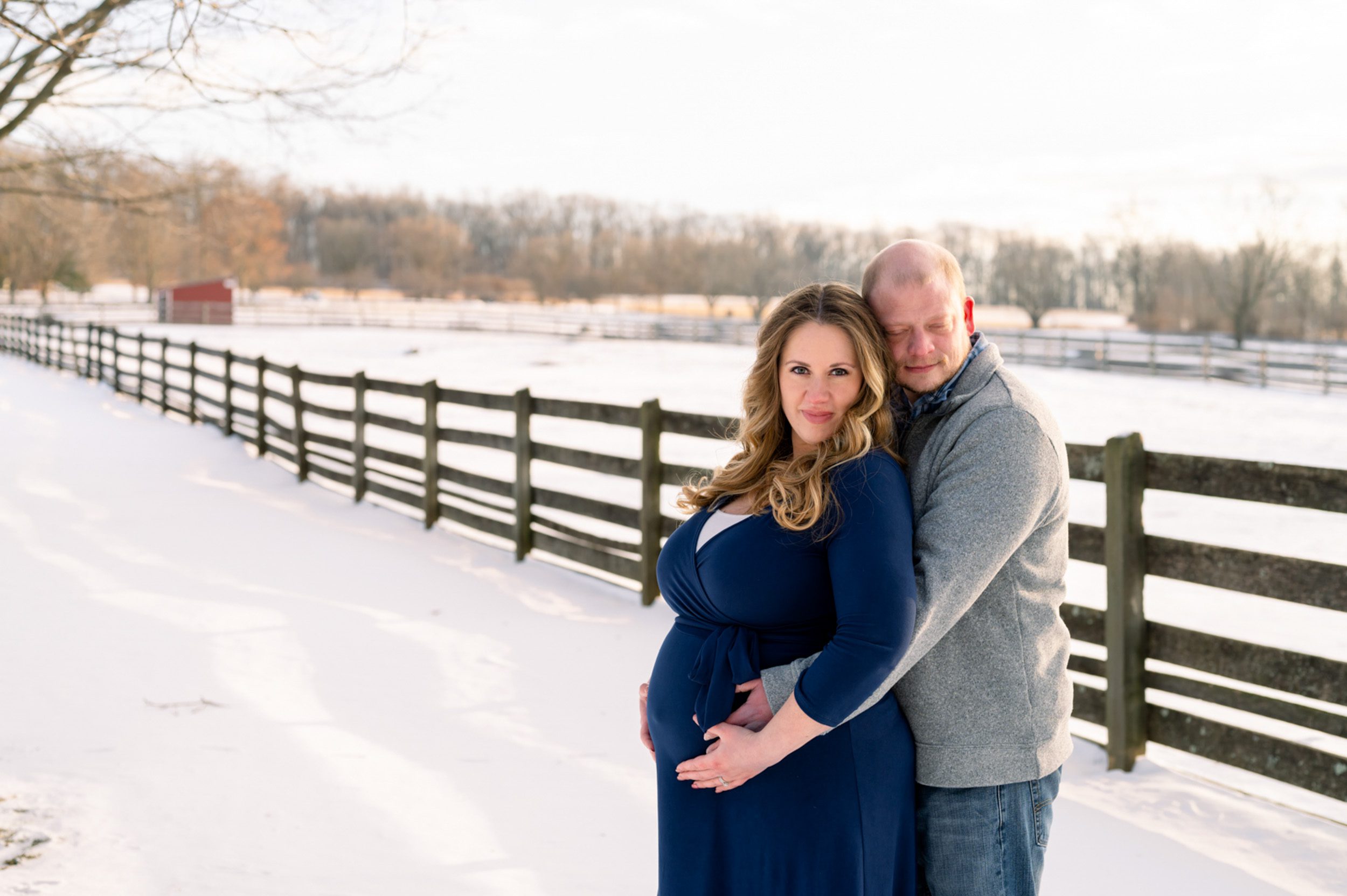 expecting mother in a navy blue dress smiling at the camera while dad hugs her from behind with his eyes closed in a snowy field at a maternity photoshoot