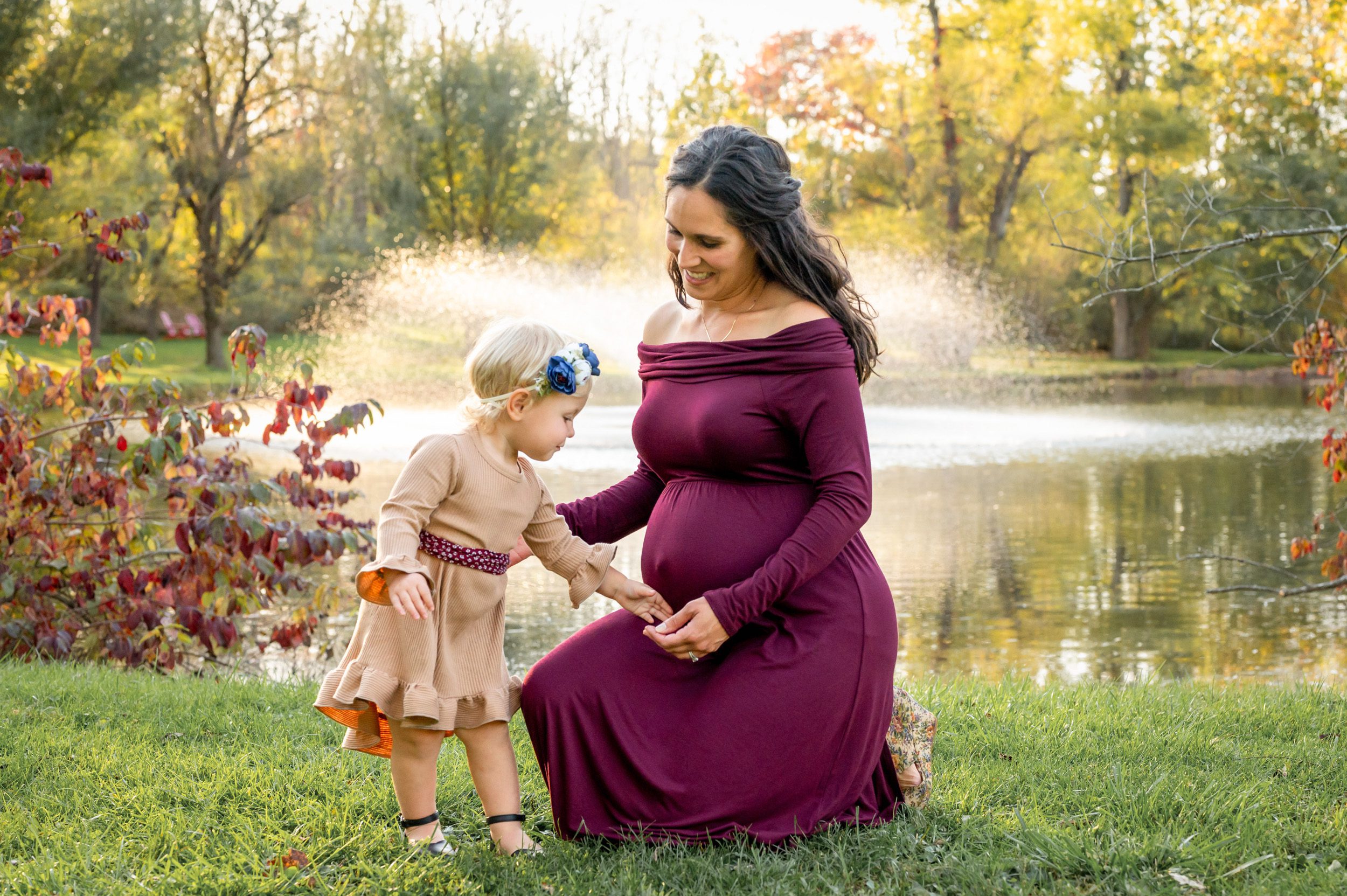 A young girl gently touching her expecting mom's belly in front of a fountain at a maternity photoshoot