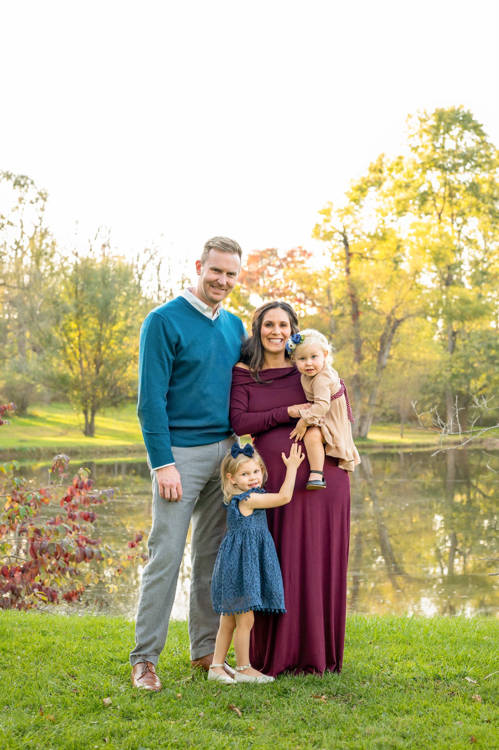 expecting parents and their two young daughters standing in front of a pond and smiling at the camera while they all touch mom's belly during a maternity photo session