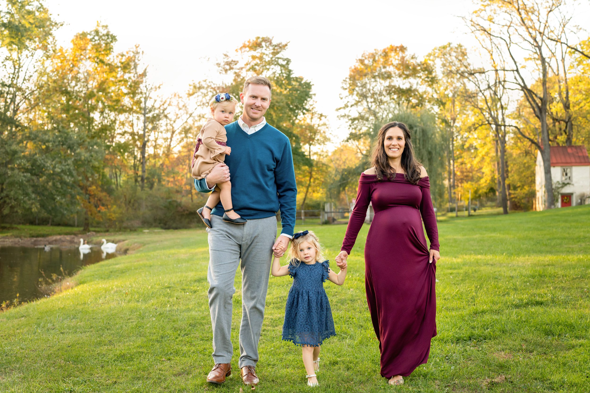 expecting parents and their two young daughters holding hands and walking through the grass next to a pond during a Phoenixville maternity photoshoot