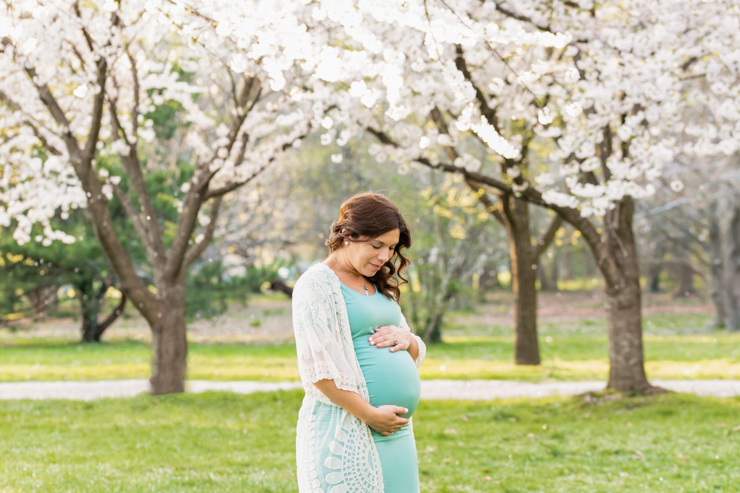 Expecting mother in an blue dress and white lace cardigan smiling down at her belly with cherry blossom trees in full bloom in the background at a maternity photoshoot