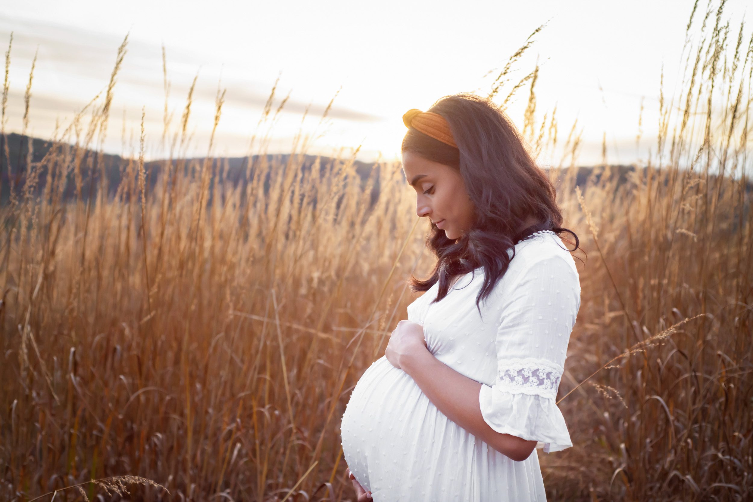 a pregnant mom in a white dress standing in a field of tall golden grasses and cradling her belly during a Valley Forge maternity photo session