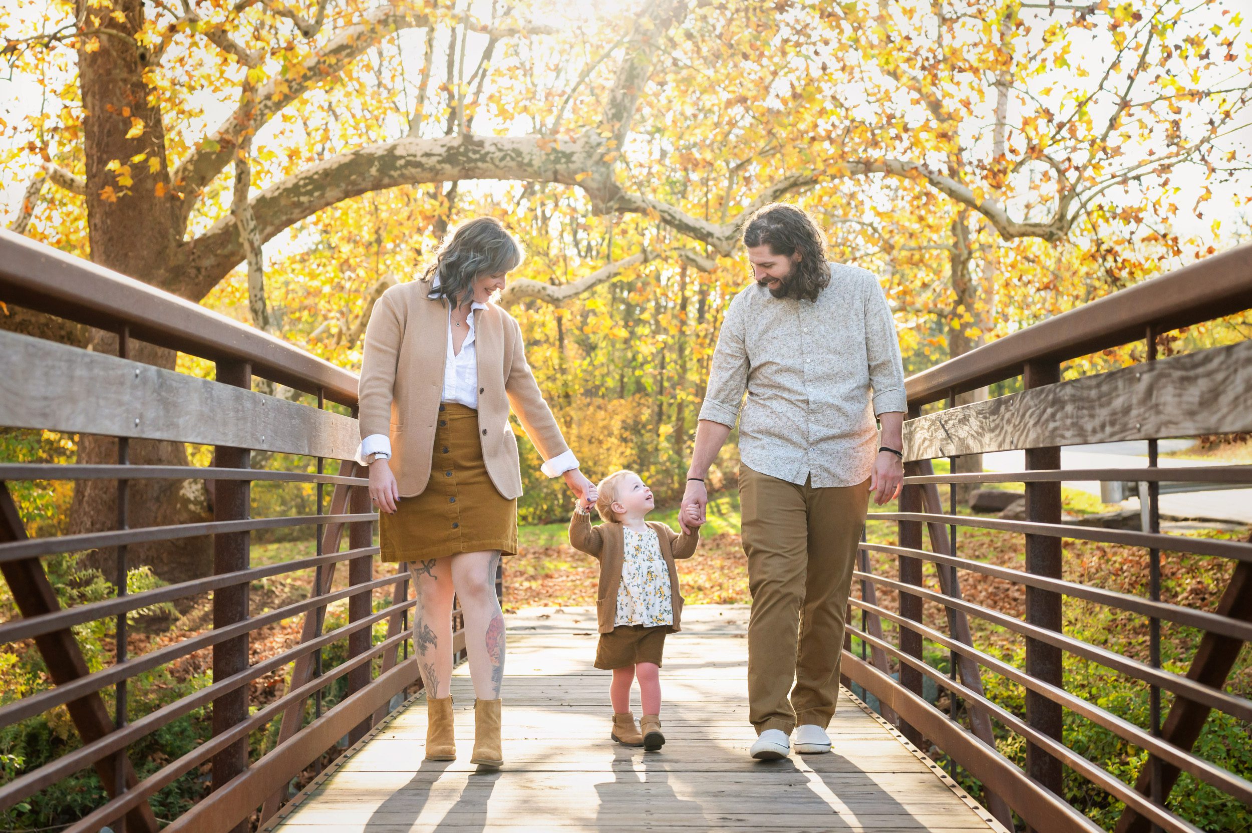 a young girl walking across a bridge holding her parents' hands and smiling up at her daddy during a Pottstown family photoshoot