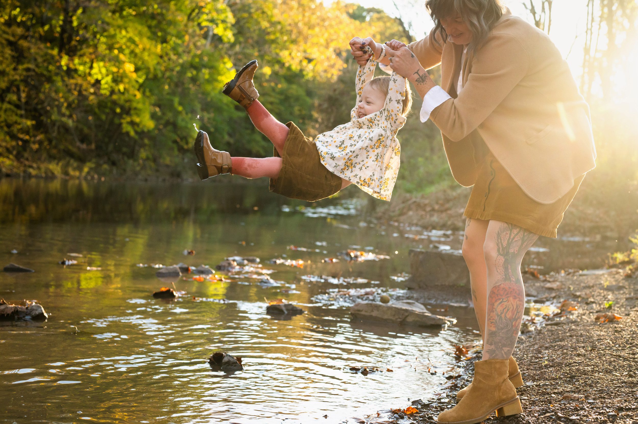 Family photo of a mom swinging her young daughter over a creek with a sun flare bursting through in the background