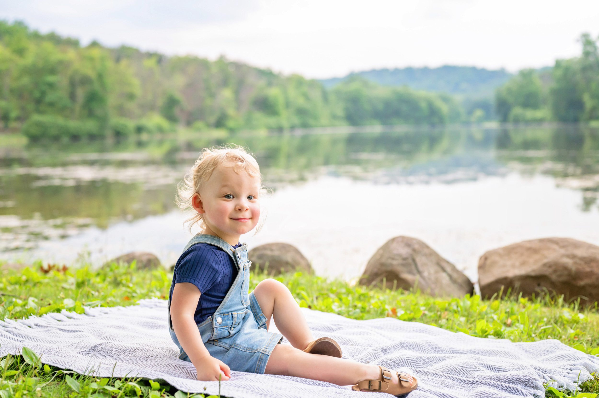 A young boy sitting on a blanket in front of a lake and smiling at the camera during a family photoshoot