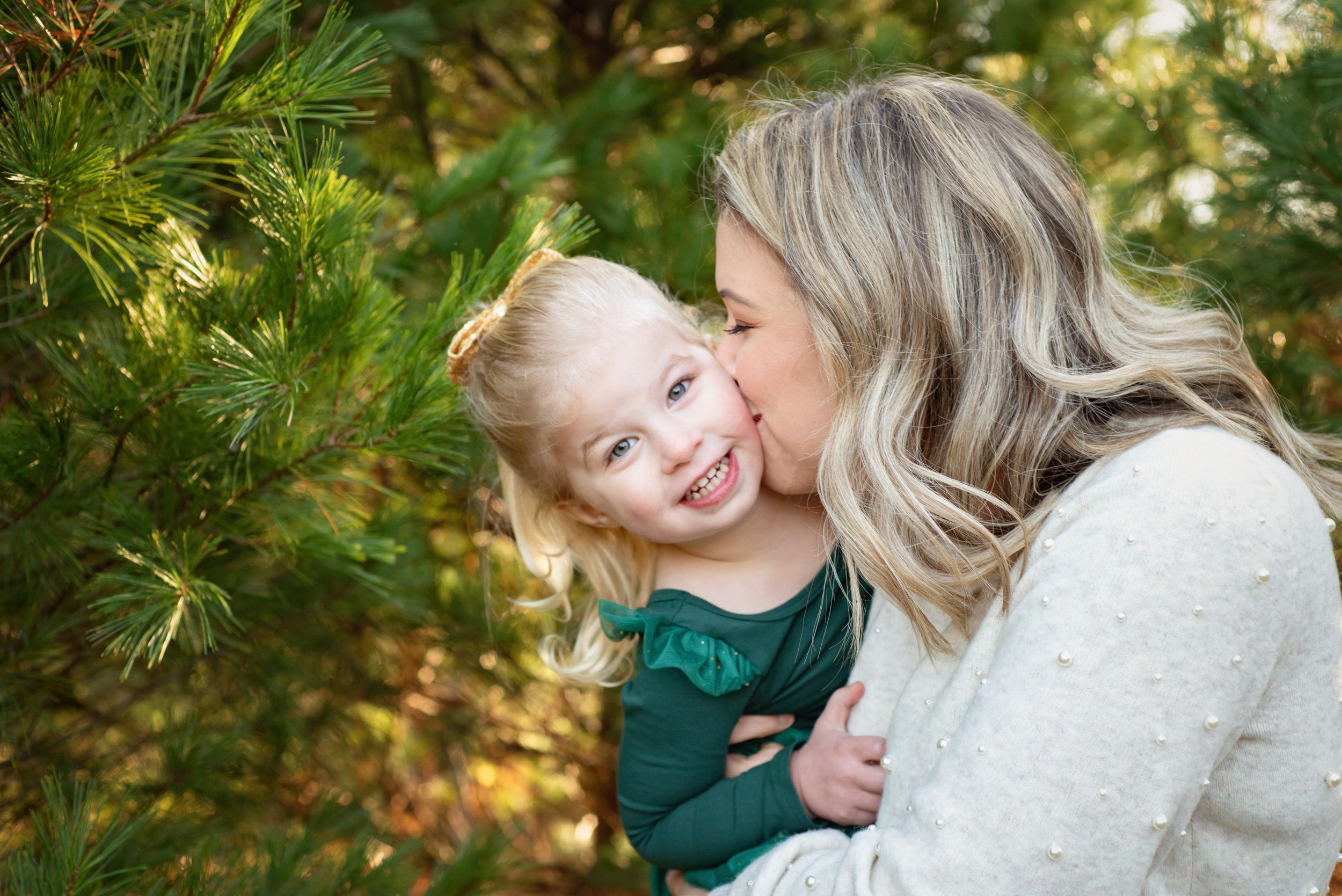 A young girl smiling at the camera while her mom kisses her cheek at a Christmas Tree Farm during a family photo session