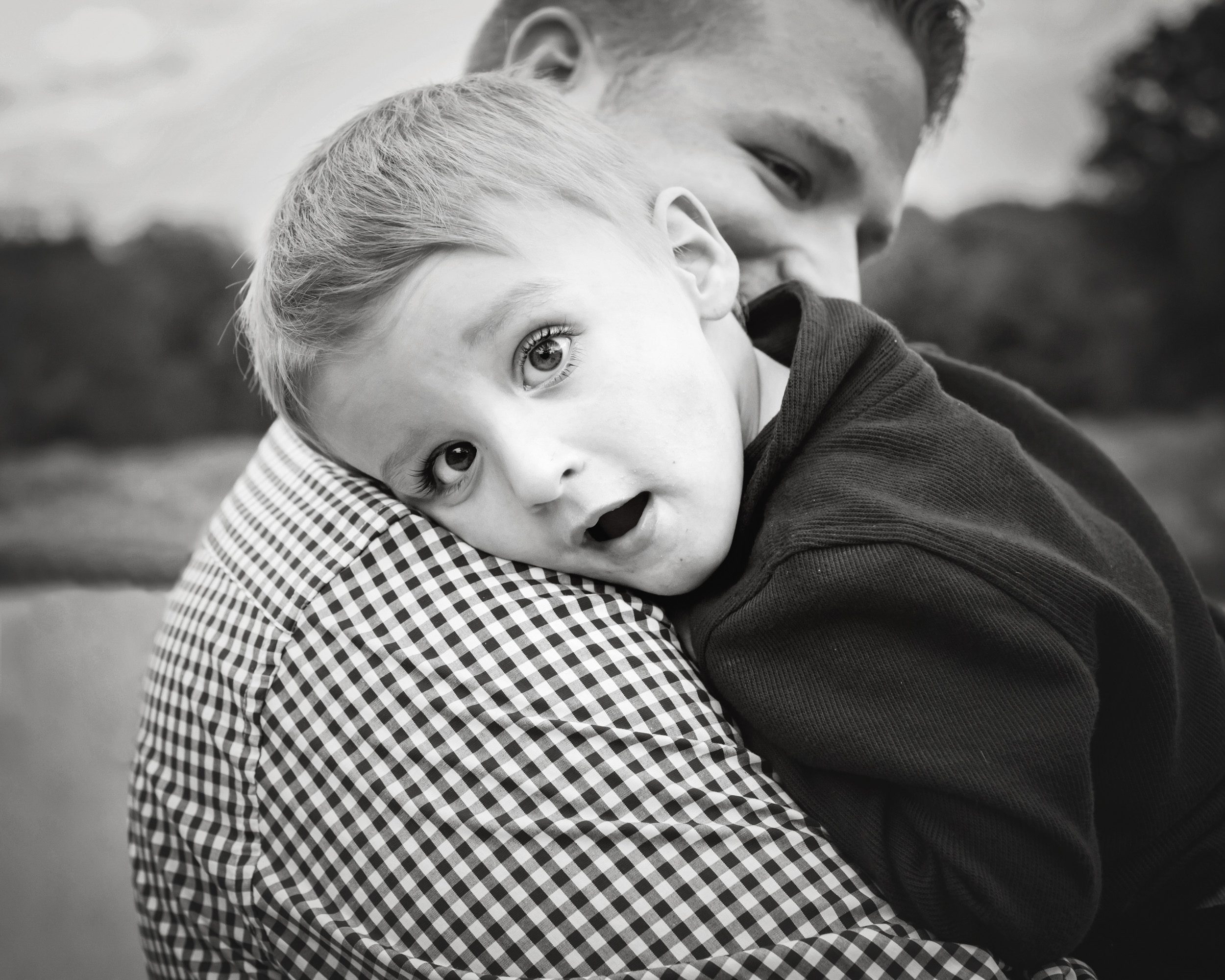 Black and white photo of a little boy resting on his dad's shoulder and gazing at the camera during a family photoshoot