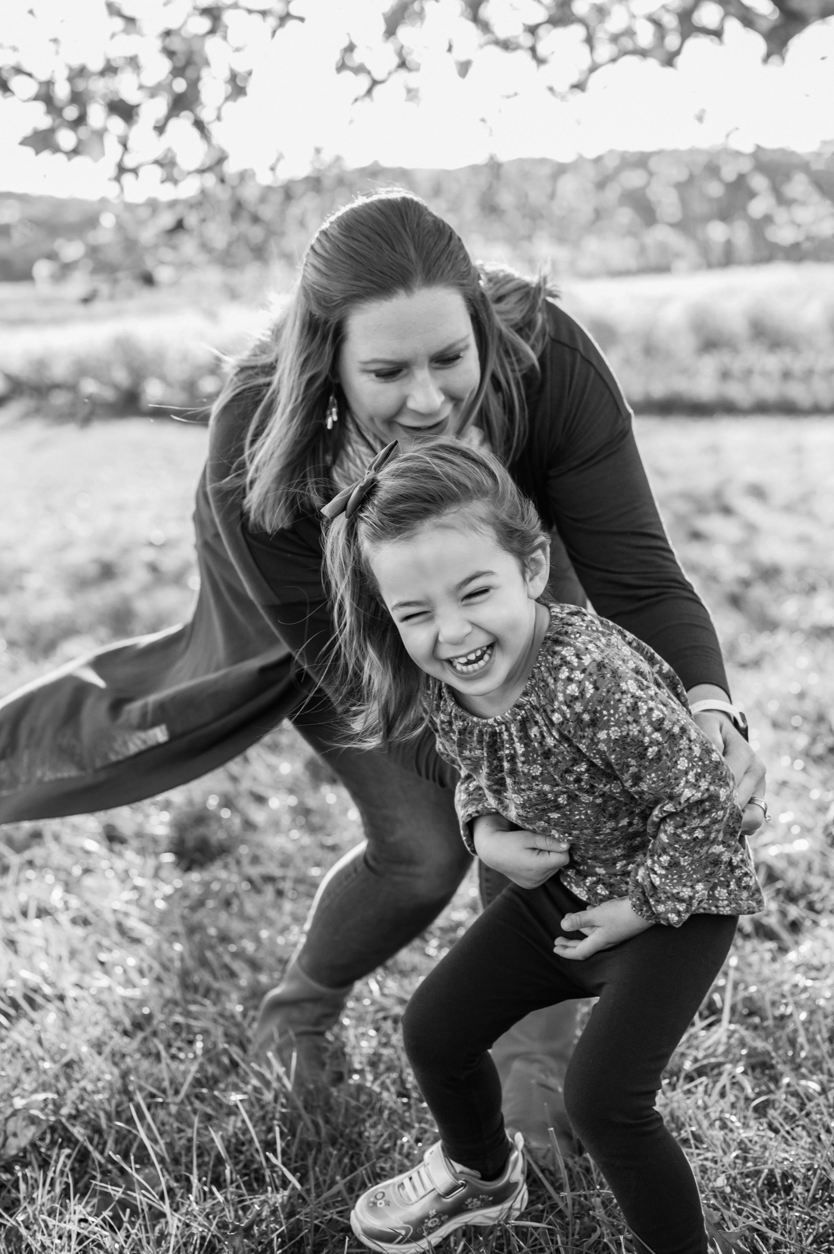 Black and white picture of a young girl laughing as her mom chases her through the grass and tickles her during a family photoshoot