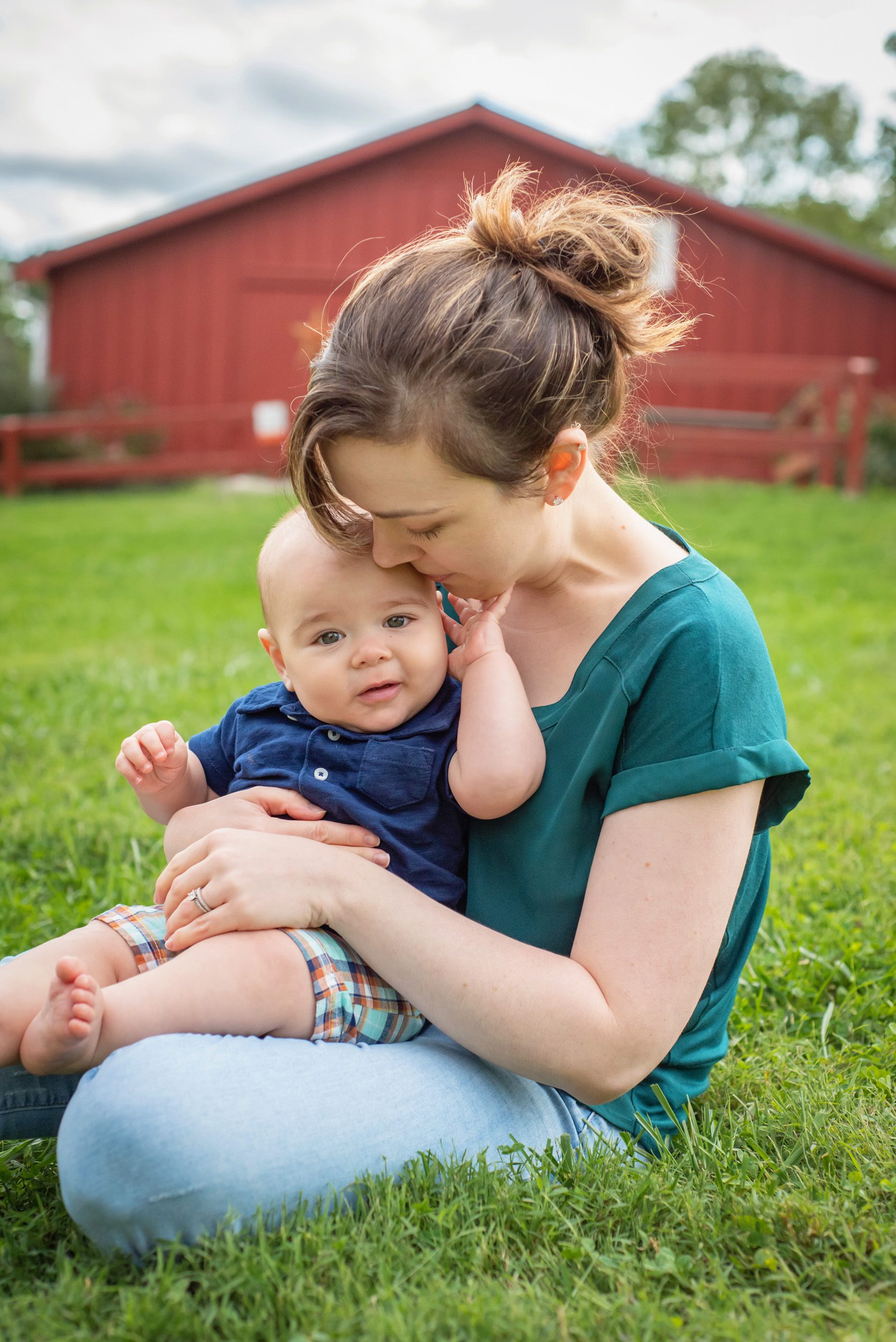 a mom hugging her young son in the grass in front of a red barn during a family photoshoot