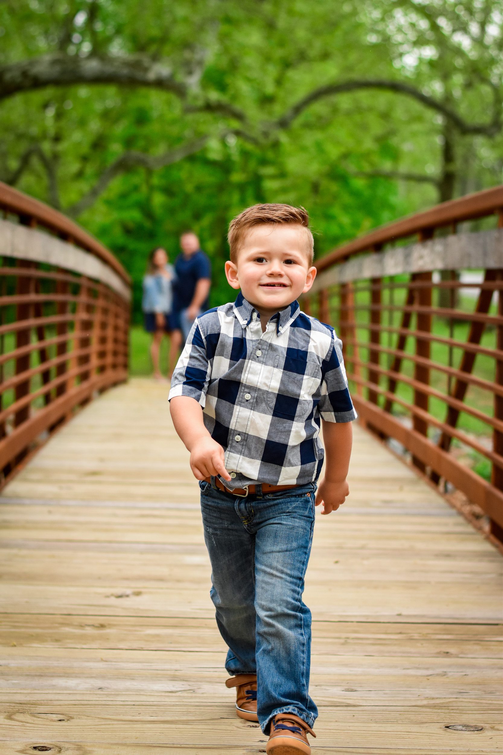 Family picture of a toddler walking across a bridge with his parents watching him in the background