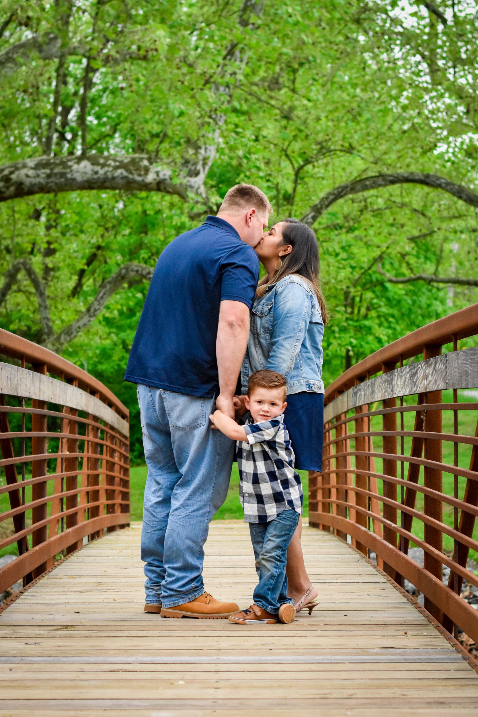 Two parents kissing on a bridge while their young son holds their hands and turns to look at the camera at a family photo session