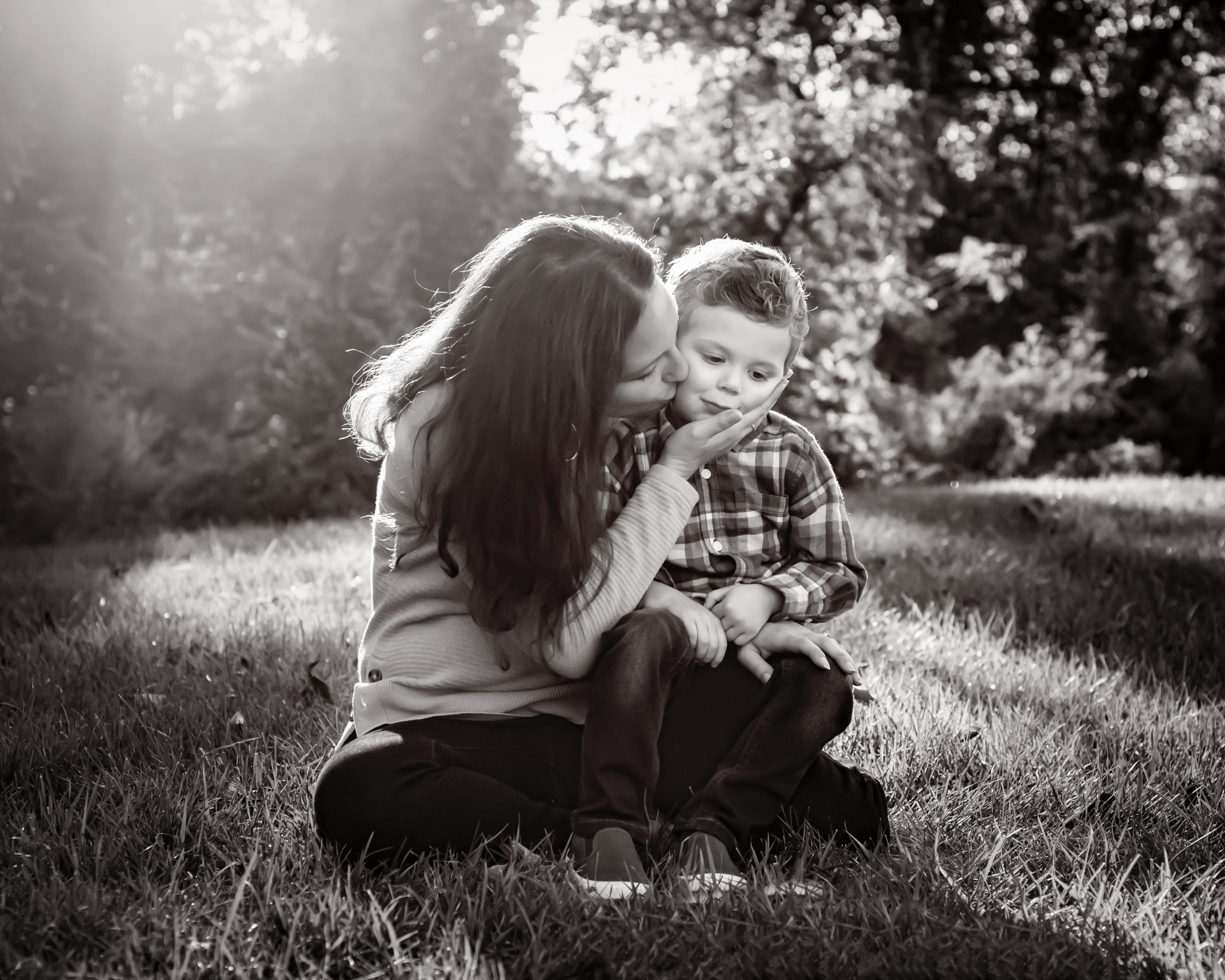 Black and white photo of a mom holding her young son on her lap and kissing him on the cheek during a family photoshoot