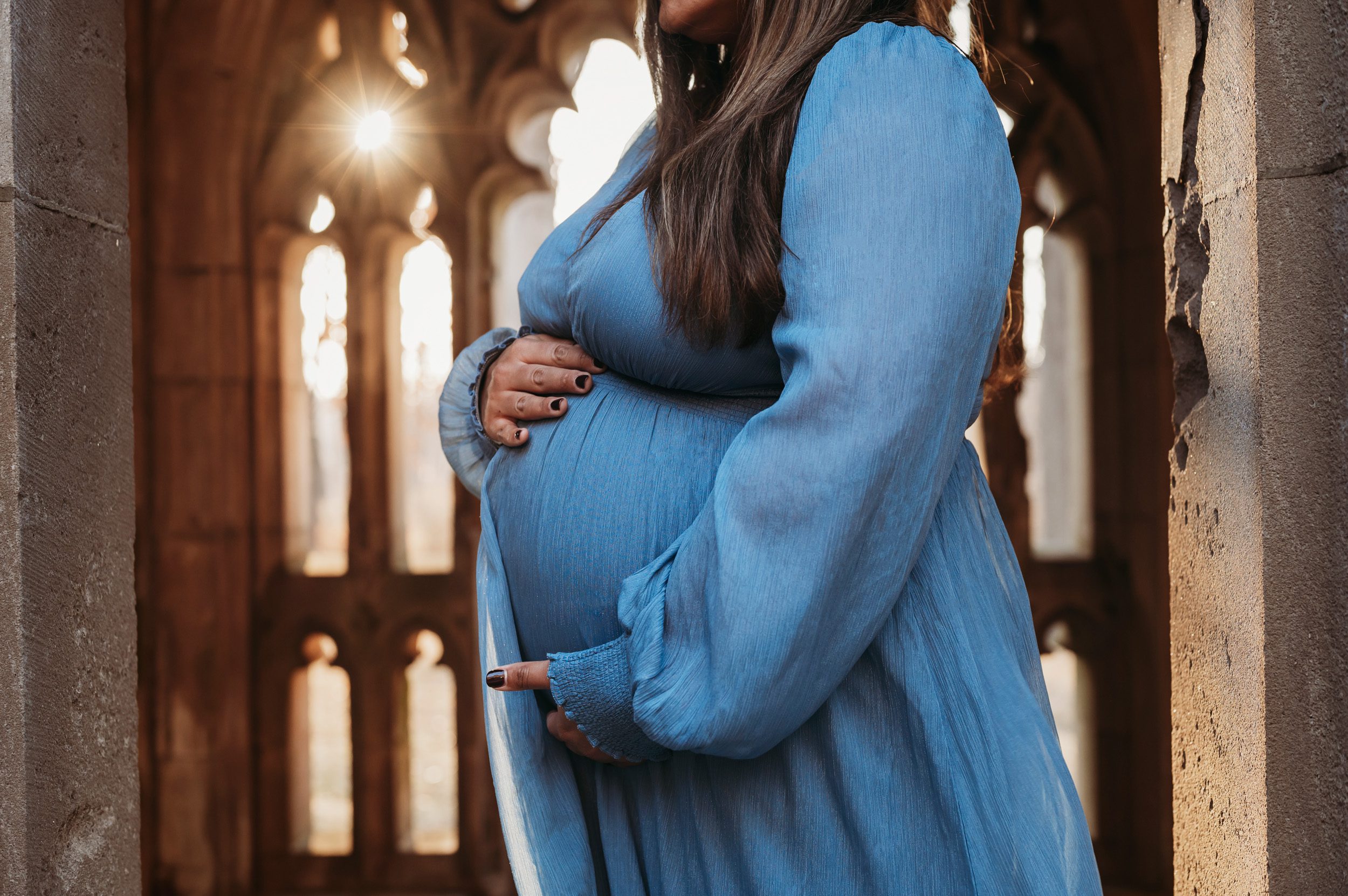 close up picture of an expecting mother in a blue dress holding her belly with sun bursting through an arch in the background at a maternity photoshoot