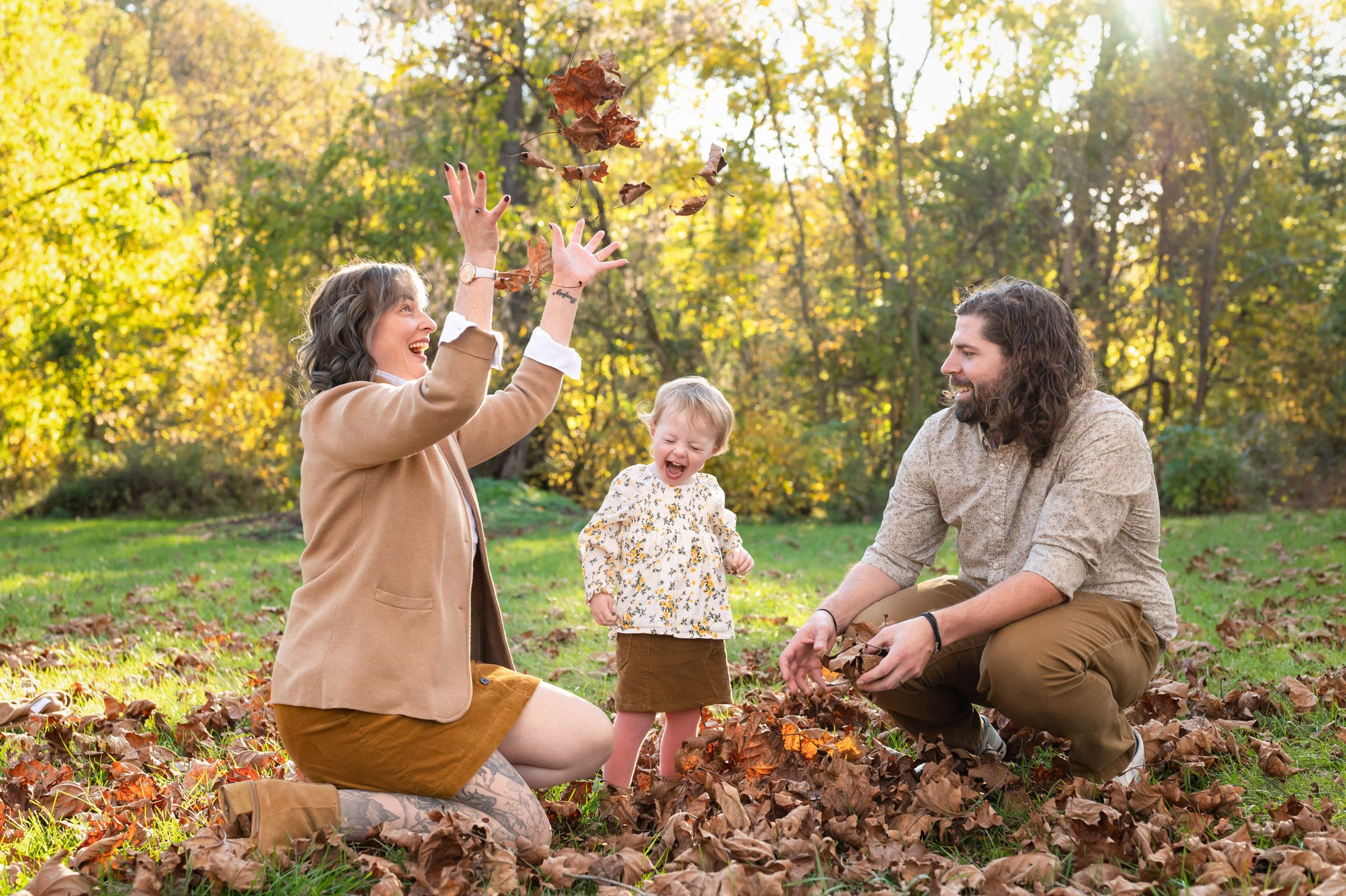 A young girl laughing in delight as her mom and dad kneel on either side of her and throw leaves up in the air during a family photoshoot