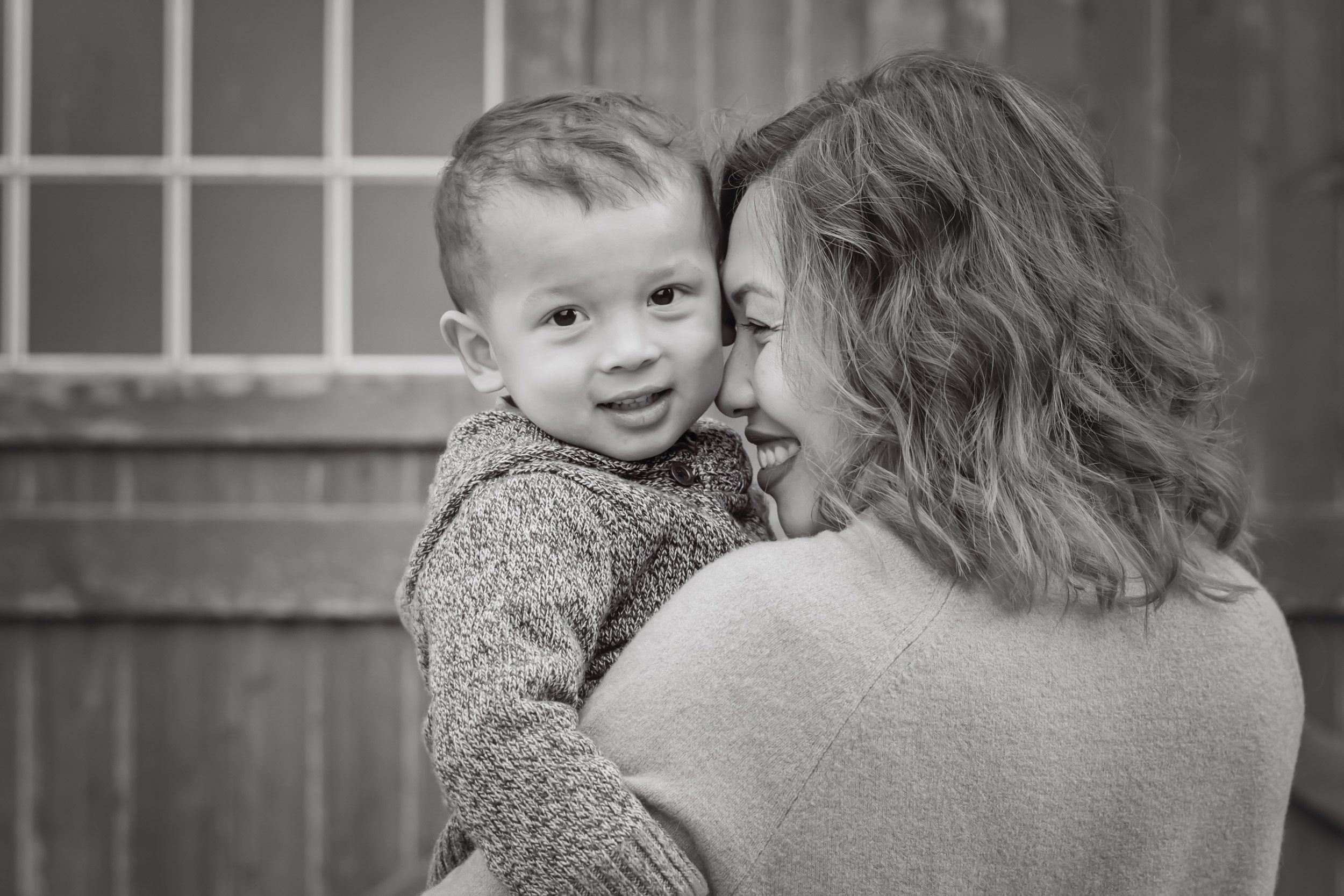 A black and white image of a little boy looking over his mom's shoulder at the camera while his mom rests her nose against his cheek and smiles at him during a family photoshoot