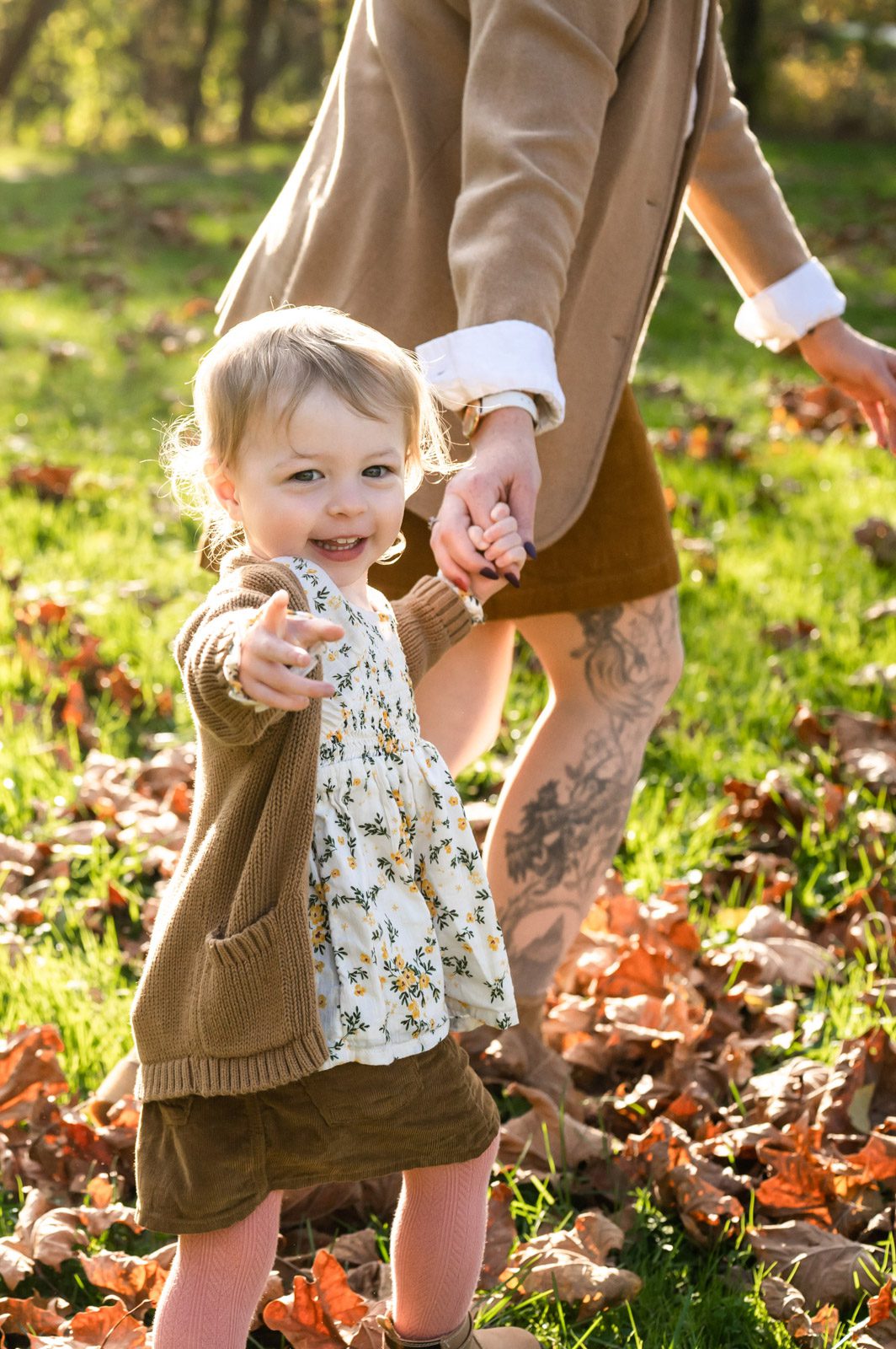 A young girl pointing at the camera and smiling as she walks through the leaves holding her mom's hand during a family photoshoot
