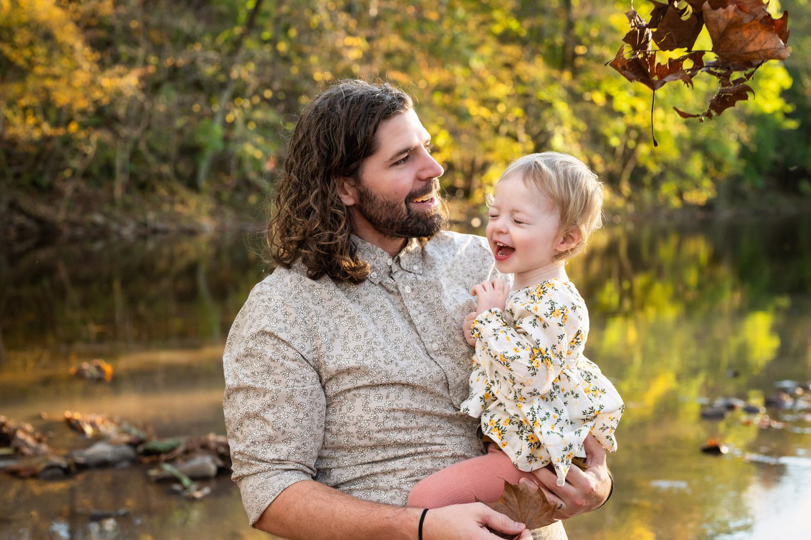 A dad holding his young daughter and laughing as mom throws leaves on them from off camera during a family photoshoot