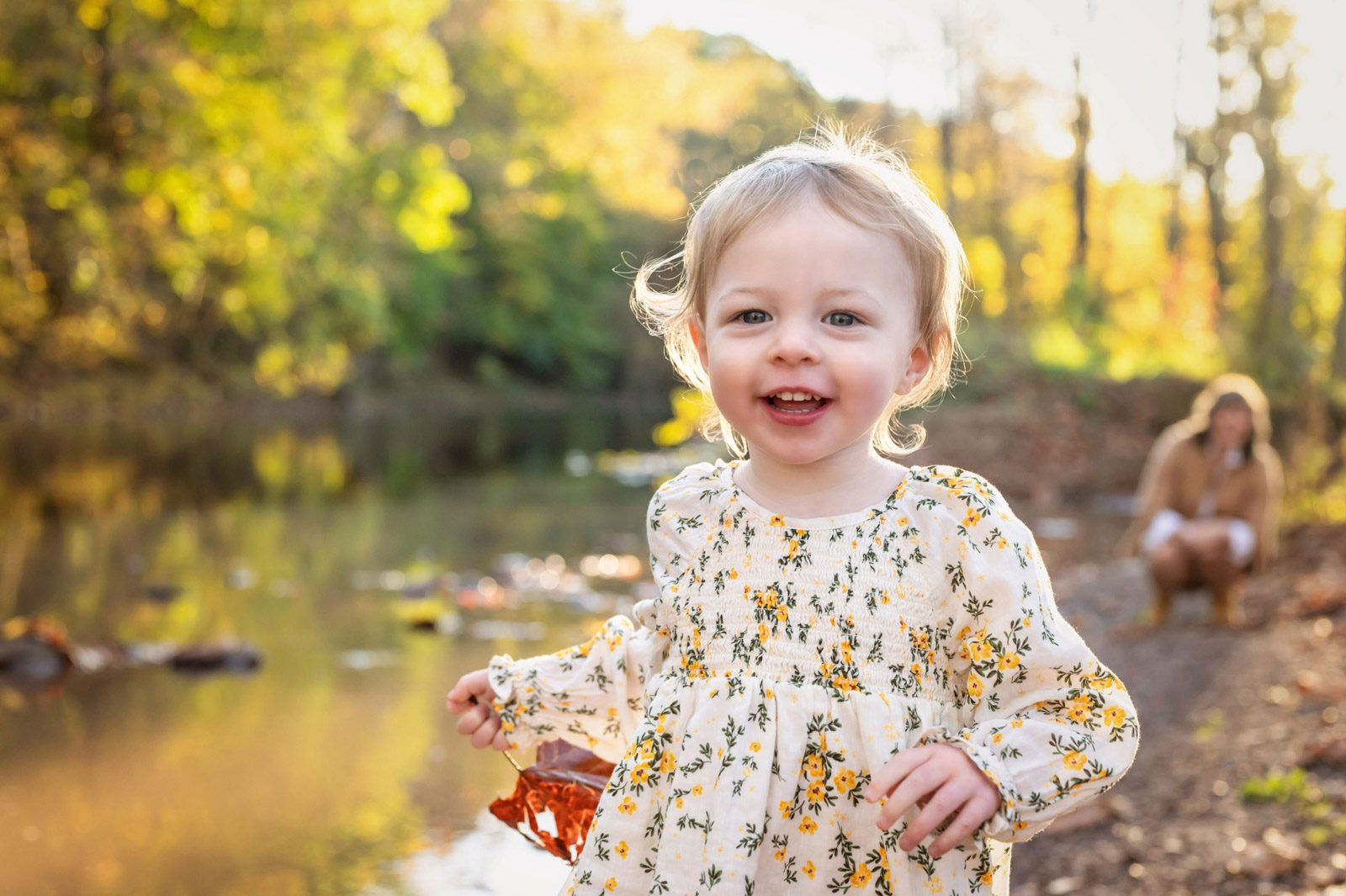 A little girl running along the edge of a creek toward the camera with her mom kneeling down and smiling at her in the background during a family photo session