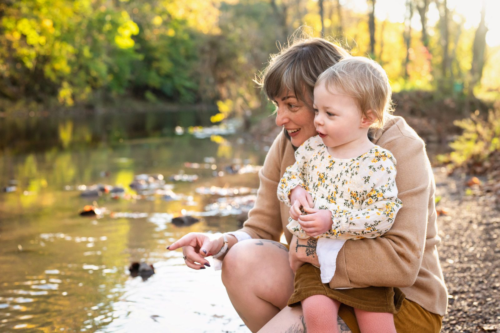 A mom kneeling next to a creek hugging her daughter and smiling as she points out a fish in the water to her daughter during a family photo session