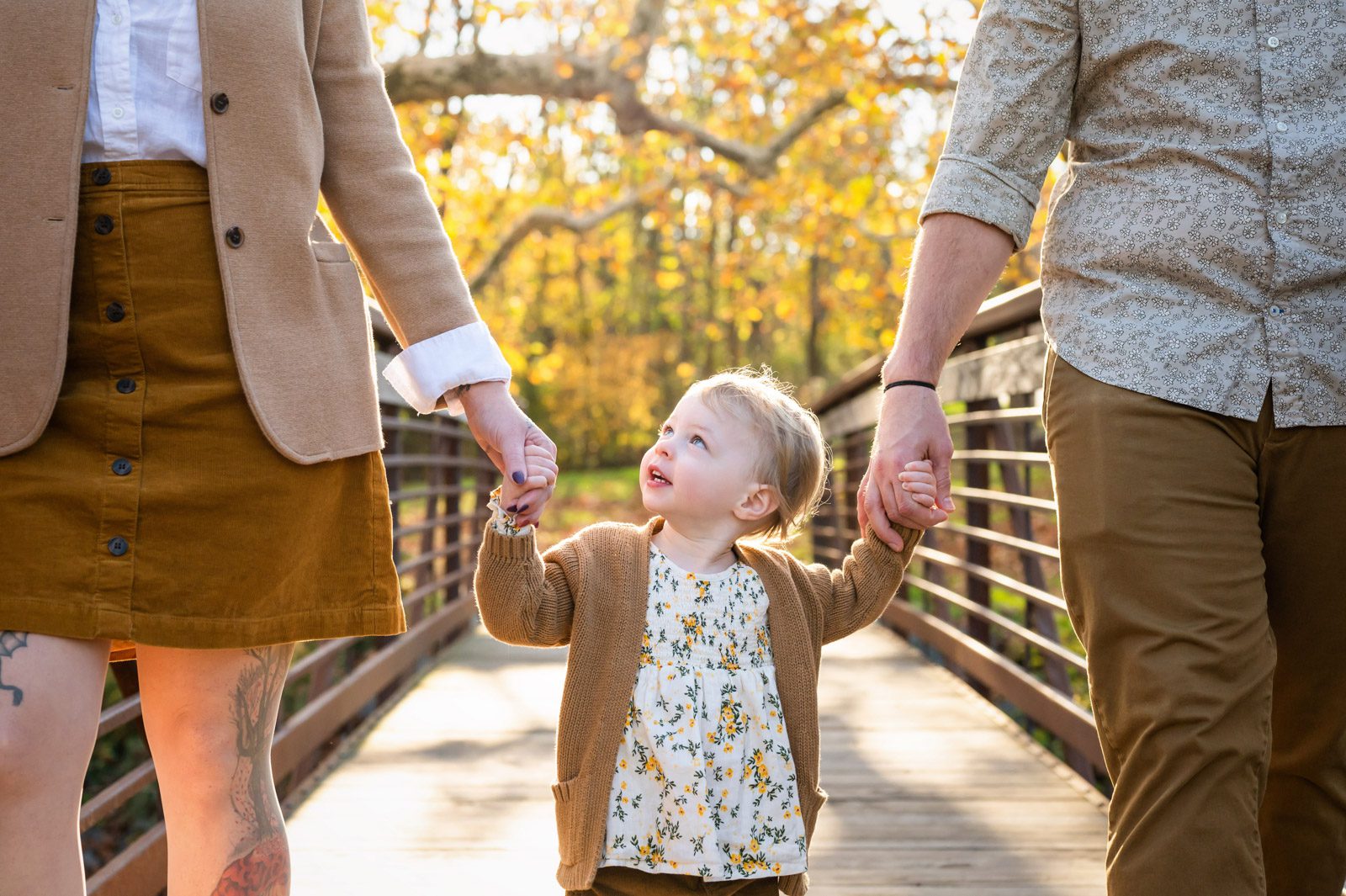 A close up of a little girl walking across a bridge holding her parents' hands and looking up at her mom during a family photoshoot