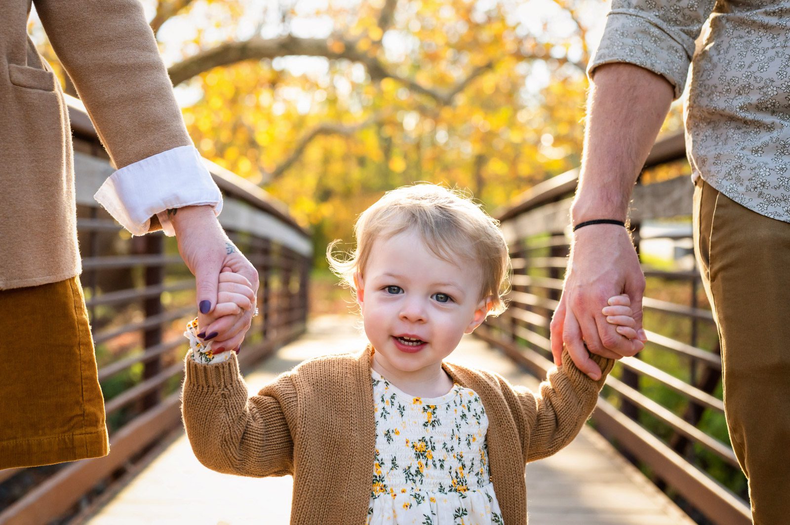 A close up image of a young girl looking at the camera and smiling as she walks across a bridge holding her parents' hands during a family photoshoot
