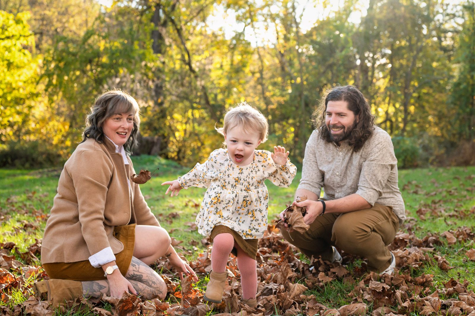 A little girl running through the leaves toward the camera with her parents kneeling in the background and watching her run during a family photoshoot