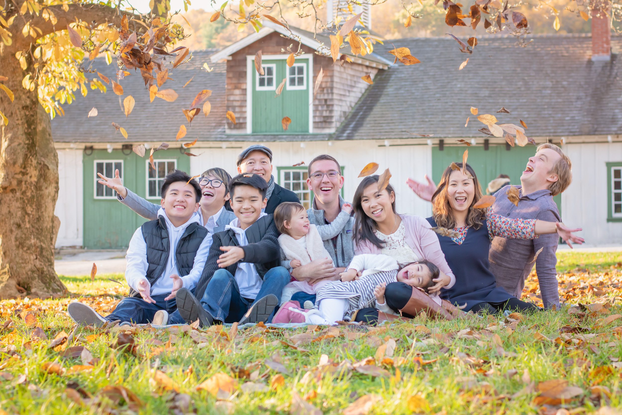extended family sitting in front of a green and white barn throwing leaves up in the air and laughing during an extended family photoshoot
