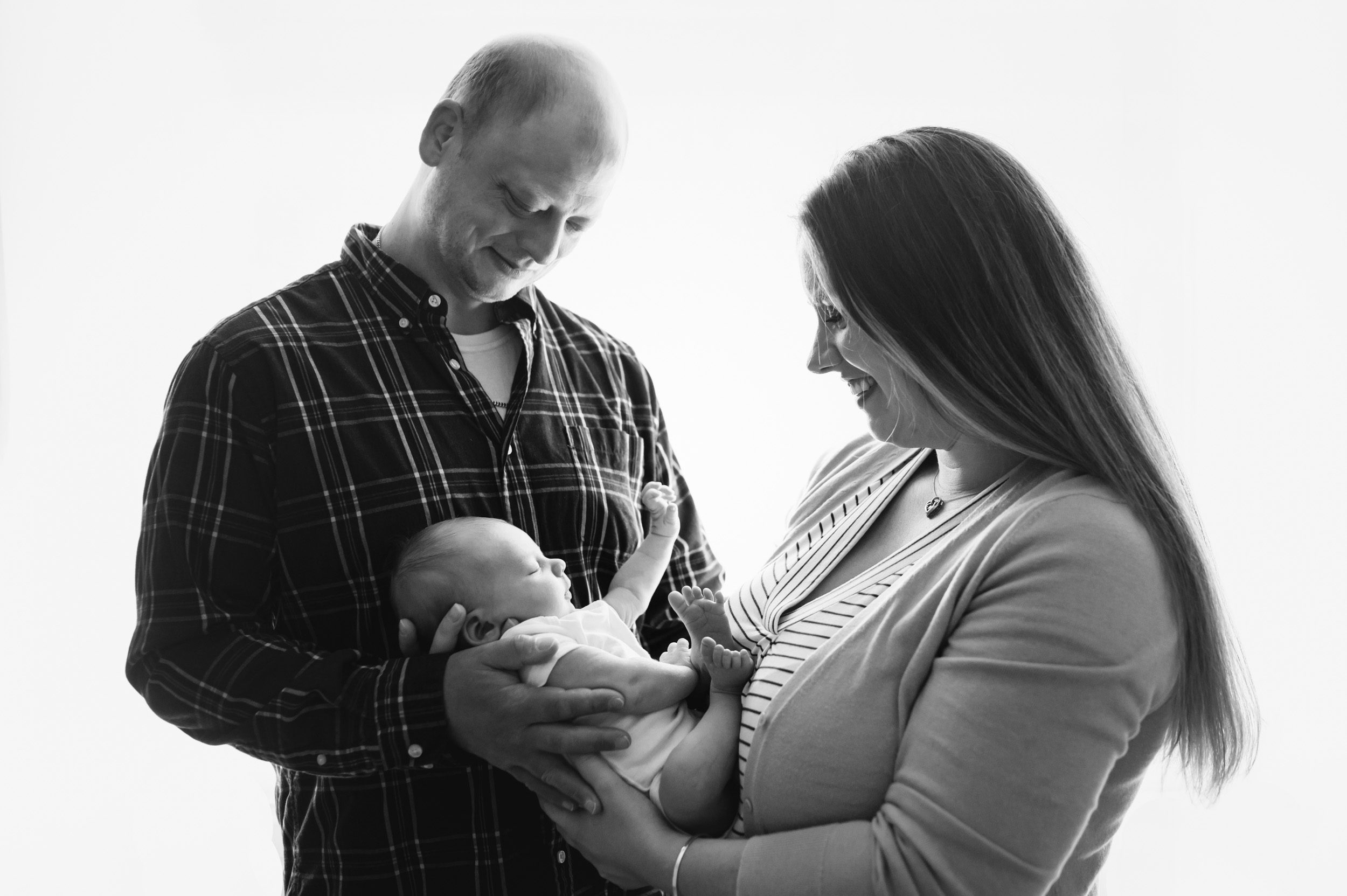 Black and white backlit image of parents holding their newborn son in their arms and smiling down at him during a natural newborn photoshoot