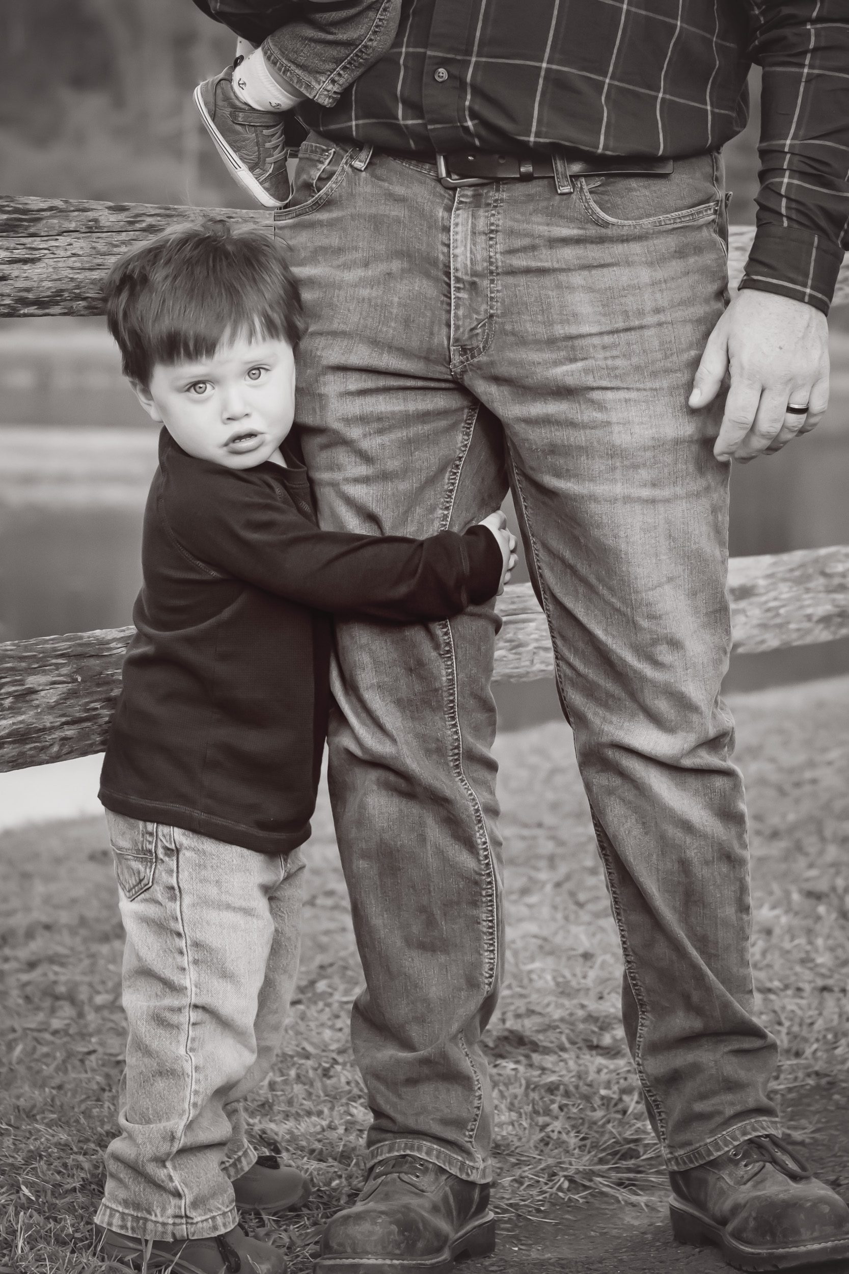 black and white image of a little boy hugging his dad's leg and looking at the camera with a pouty face at a family photoshoot