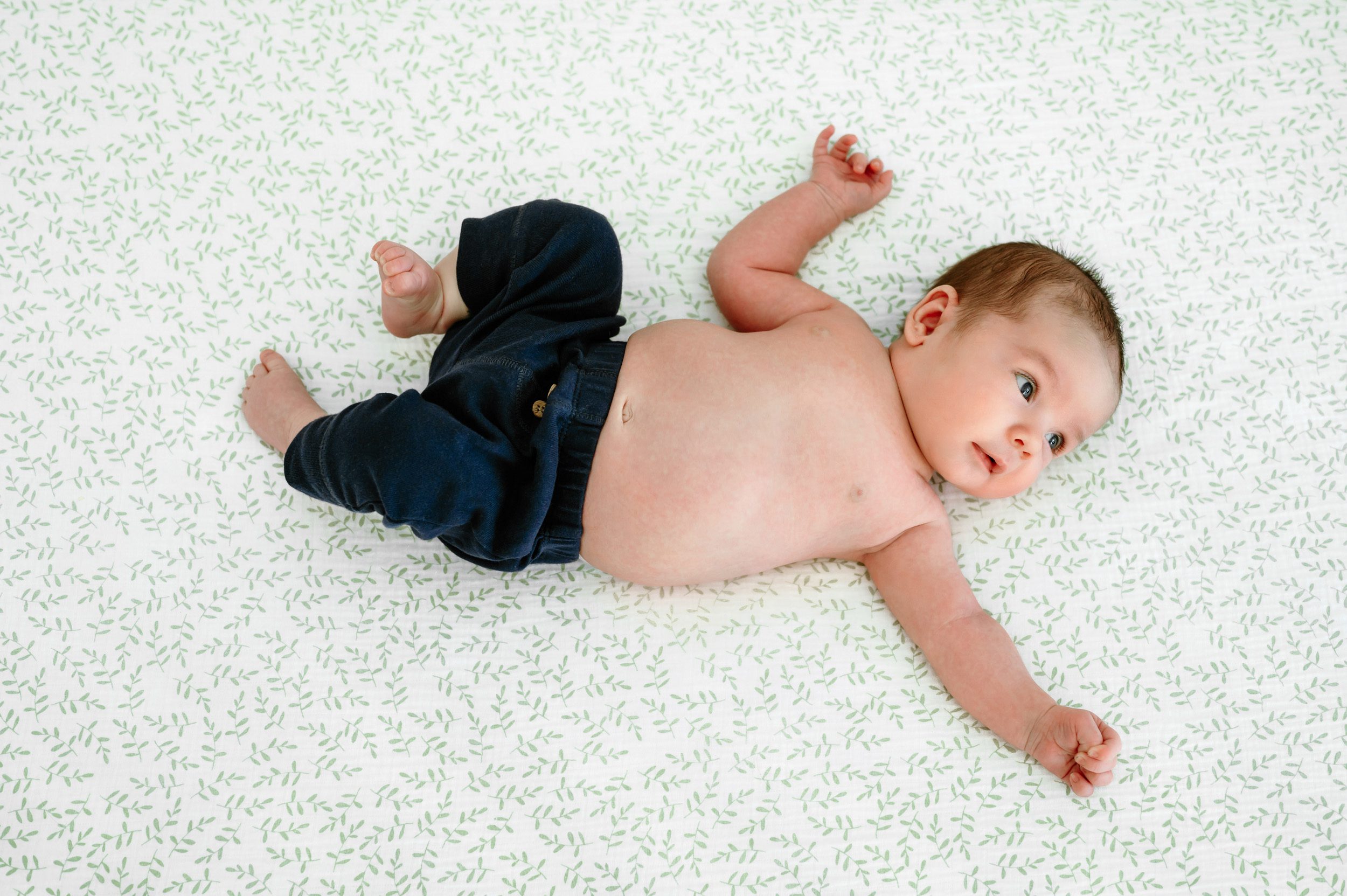 An older newborn baby laying in his crib on a white crib mattress with green leaves and reaching above his head toward the light of the window during a newborn photo session