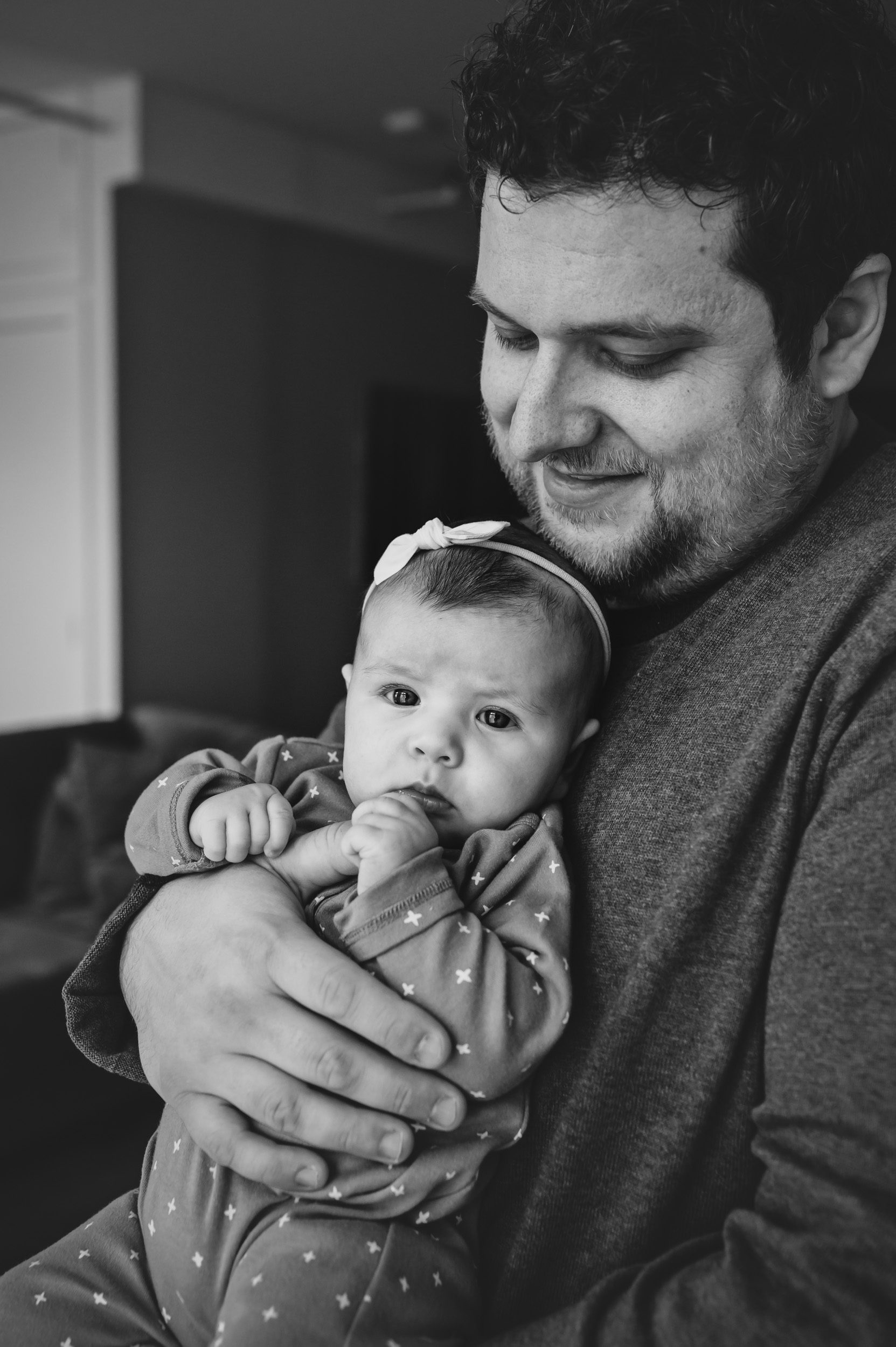 Black and white image of a new father holding his baby girl and smiling down at her as she grabs onto his thumb during a home newborn photo session