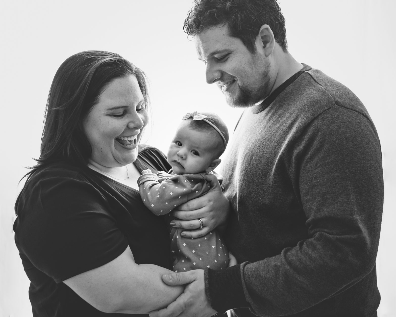 Black and white backlit image of new parents holding their baby girl in between them and smiling at her during a home newborn photo session