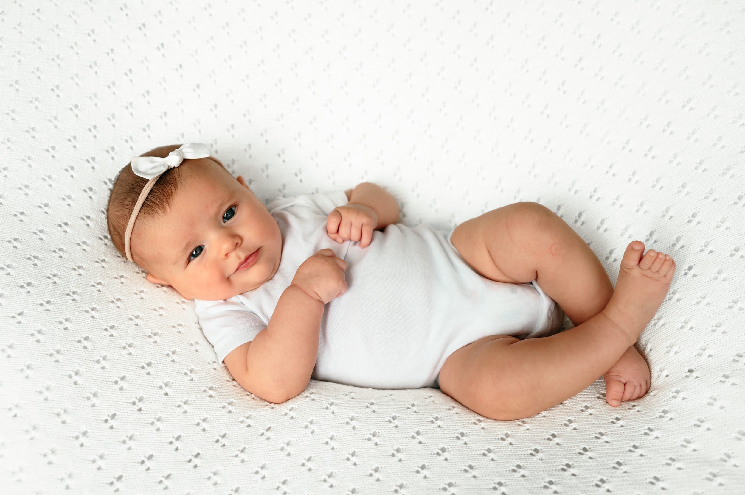 A baby girl in white onesie on a white textured backdrop smirking up at the camera during a home newborn photoshoot