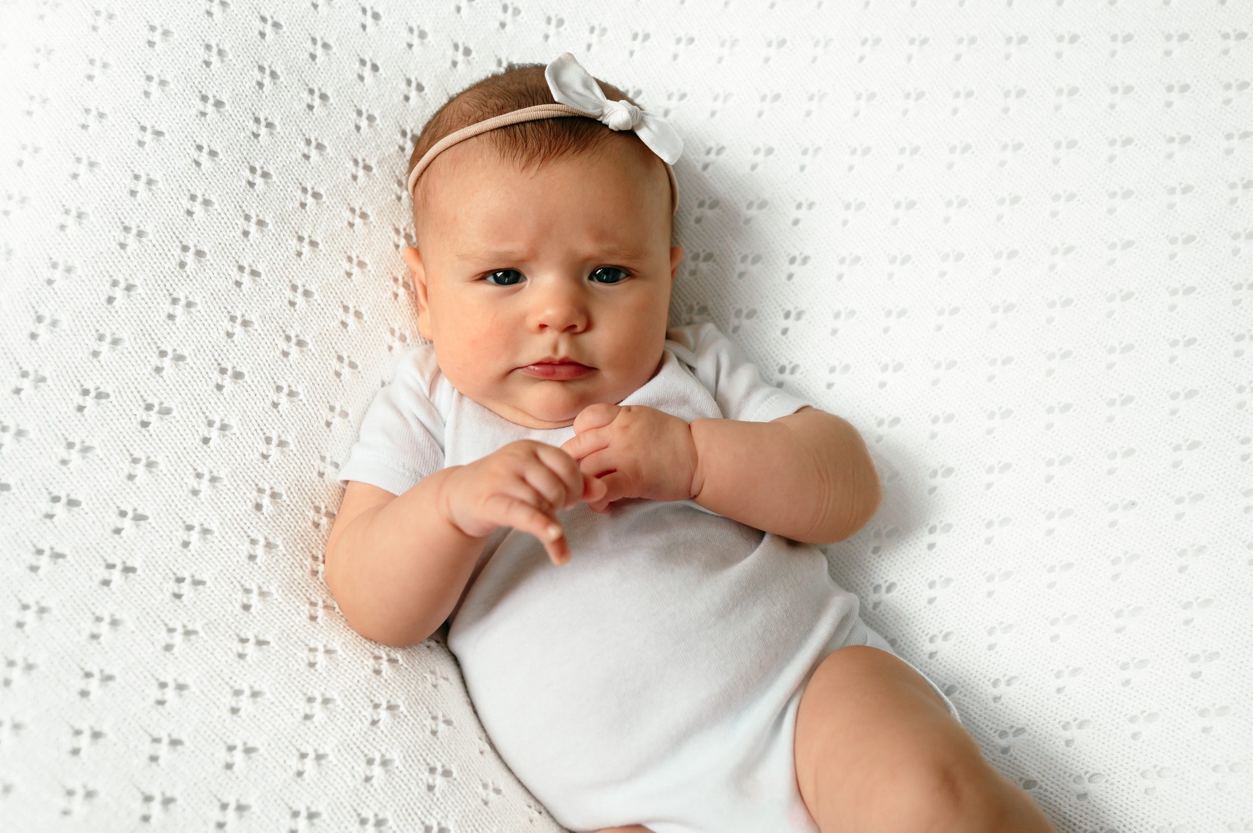 A baby girl in a white onesie on a white textured backdrop looking up at the camera with a concerned look on her face during a home newborn photoshoot