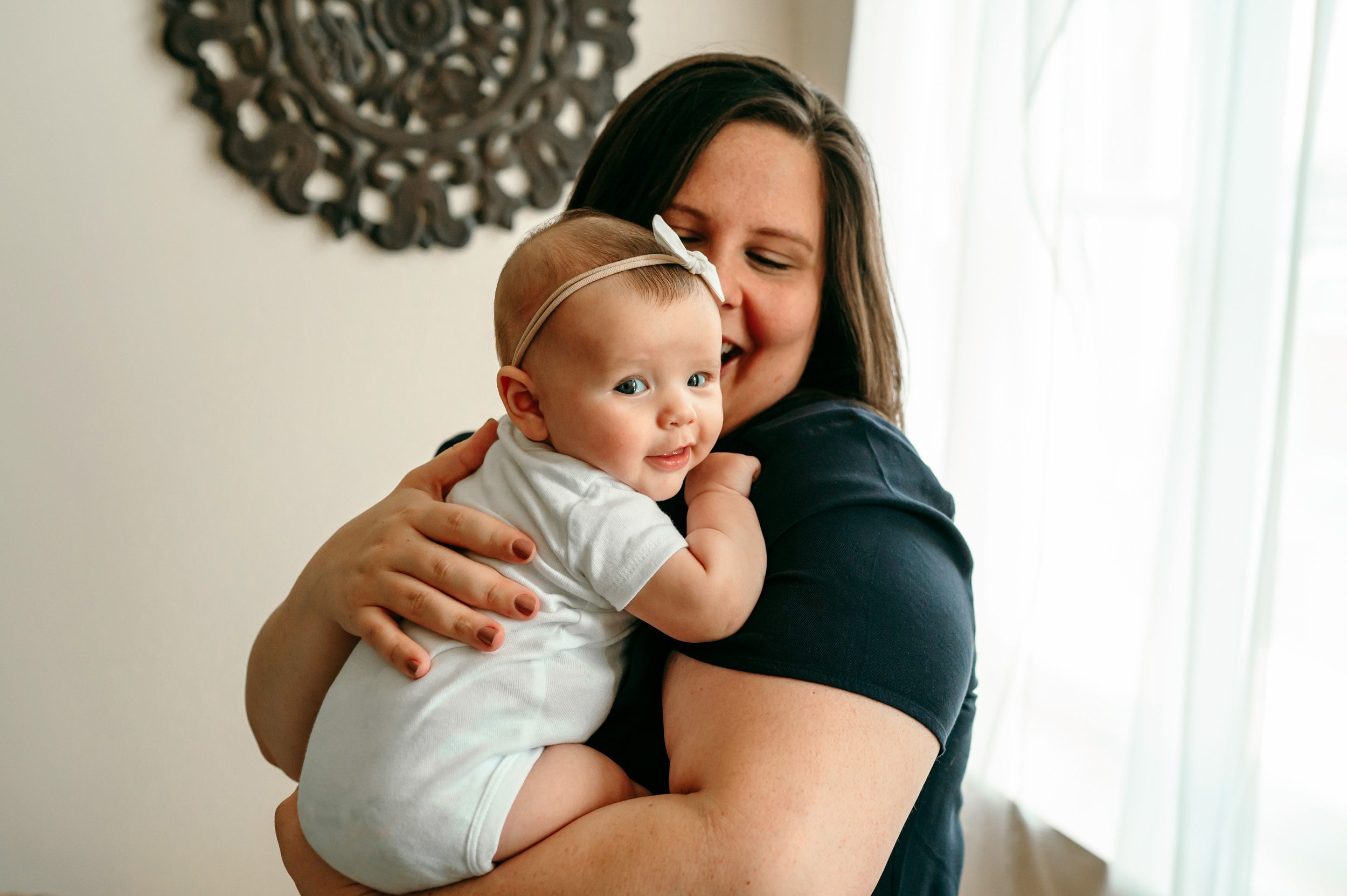 A baby girl looking back toward the camera and smiling while her mom holds her in her arms during a home newborn photoshoot