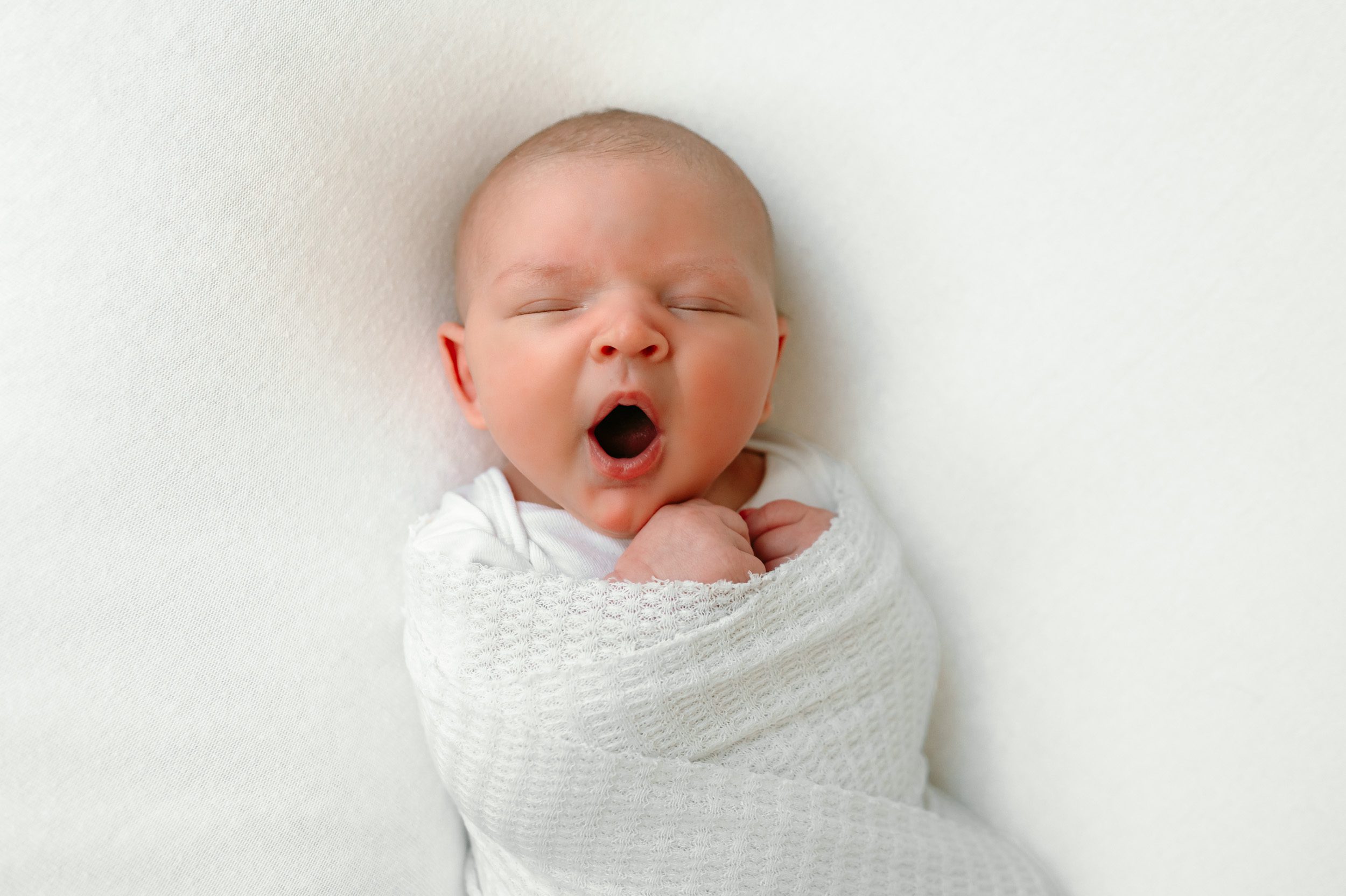 Yawning newborn baby boy in a white swaddle on a white backdrop at a home newborn photoshoot