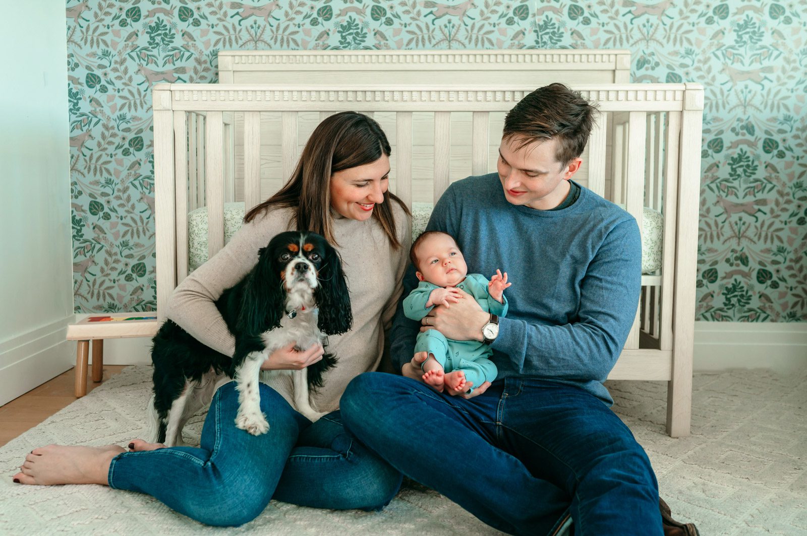 New parents sitting in front of the crib with their newborn baby boy and pet dog on their lap during a home newborn photoshoot
